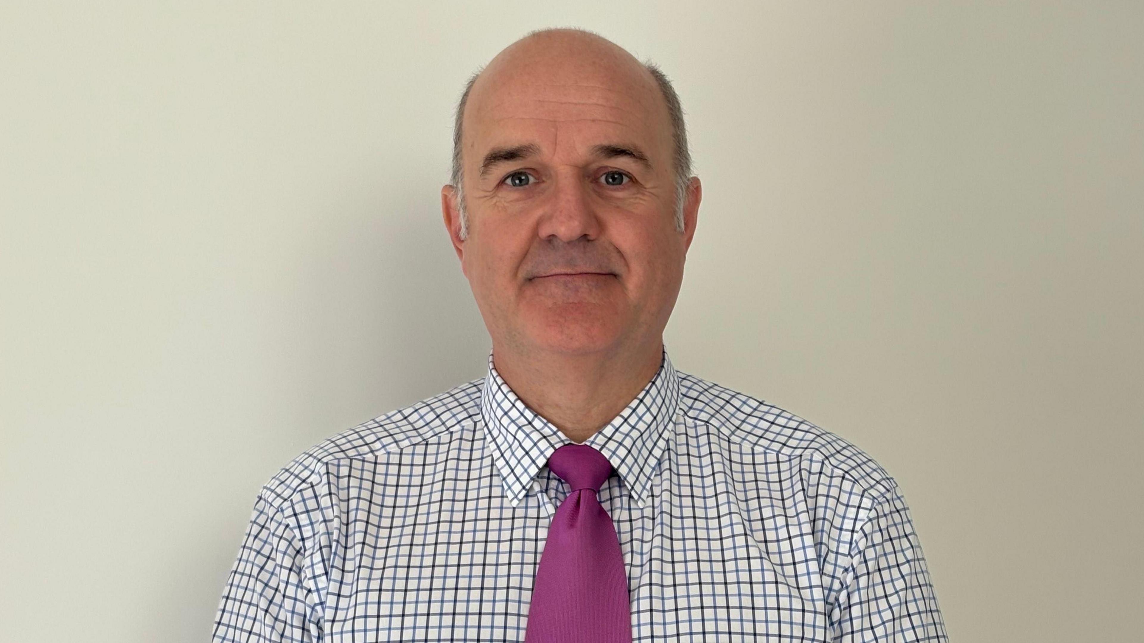 A bald man with grey hair on either side of his head wearing a white shirt with blue and red checked lines and a magenta tie standing in front of a plain white wall.