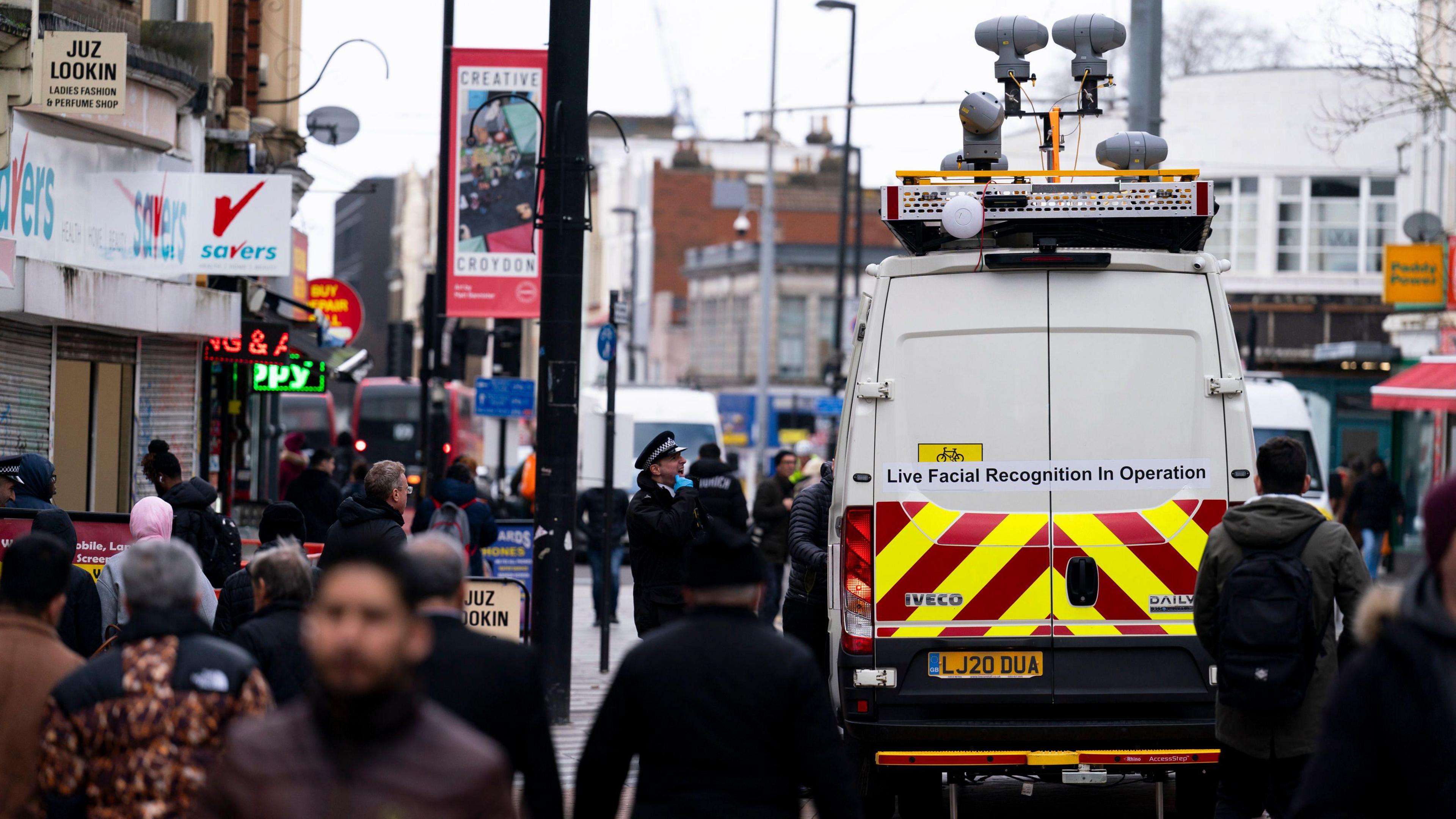 Live facial recognition cameras on top of a van in Croydon. Officers are standing close to the van, and people walk by in coats.