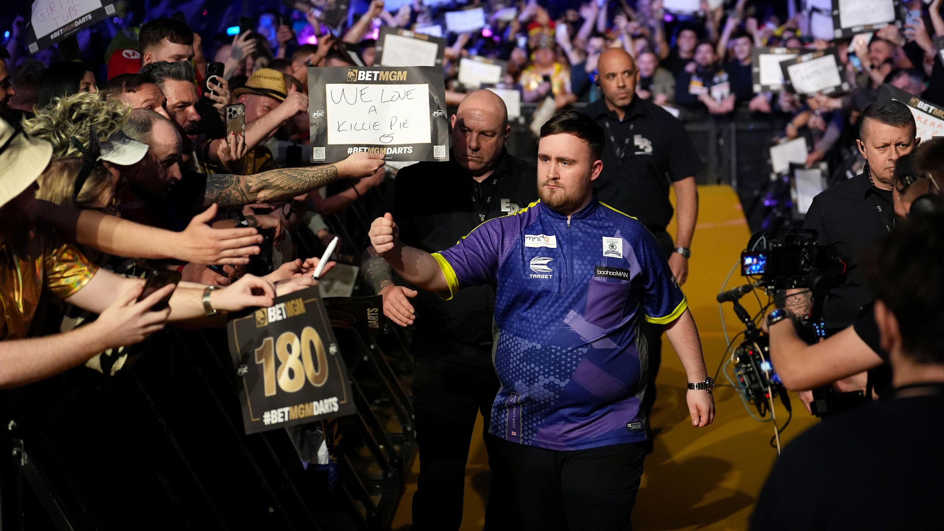 Luke Littler enters an arena surrounded by large crowds of excited fans waving placards and filmed by a camera operator