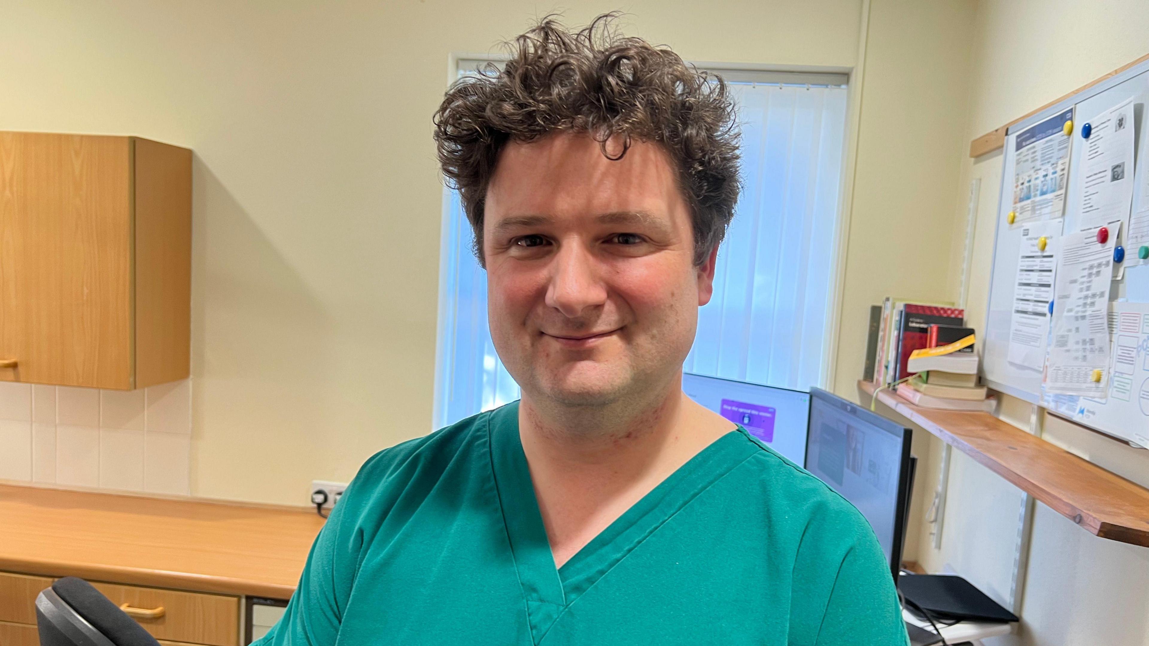 A GP with brown curly hair wearing blue overalls is standing inside a GP clinic and smiling at the camera. Behind him is a wooden table, office chair, computer and whiteboard with several pieces of paper attached to it. 