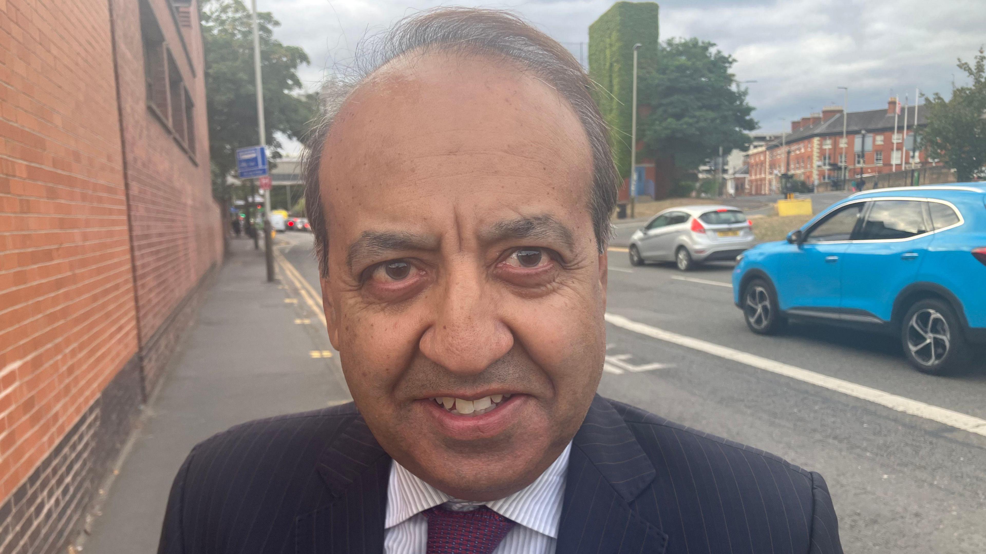 Evington councillor Zuffar Haq standing in a street in Leicester. He is a middle-aged South-Asian man wearing a dark blue suit with pinstripes.