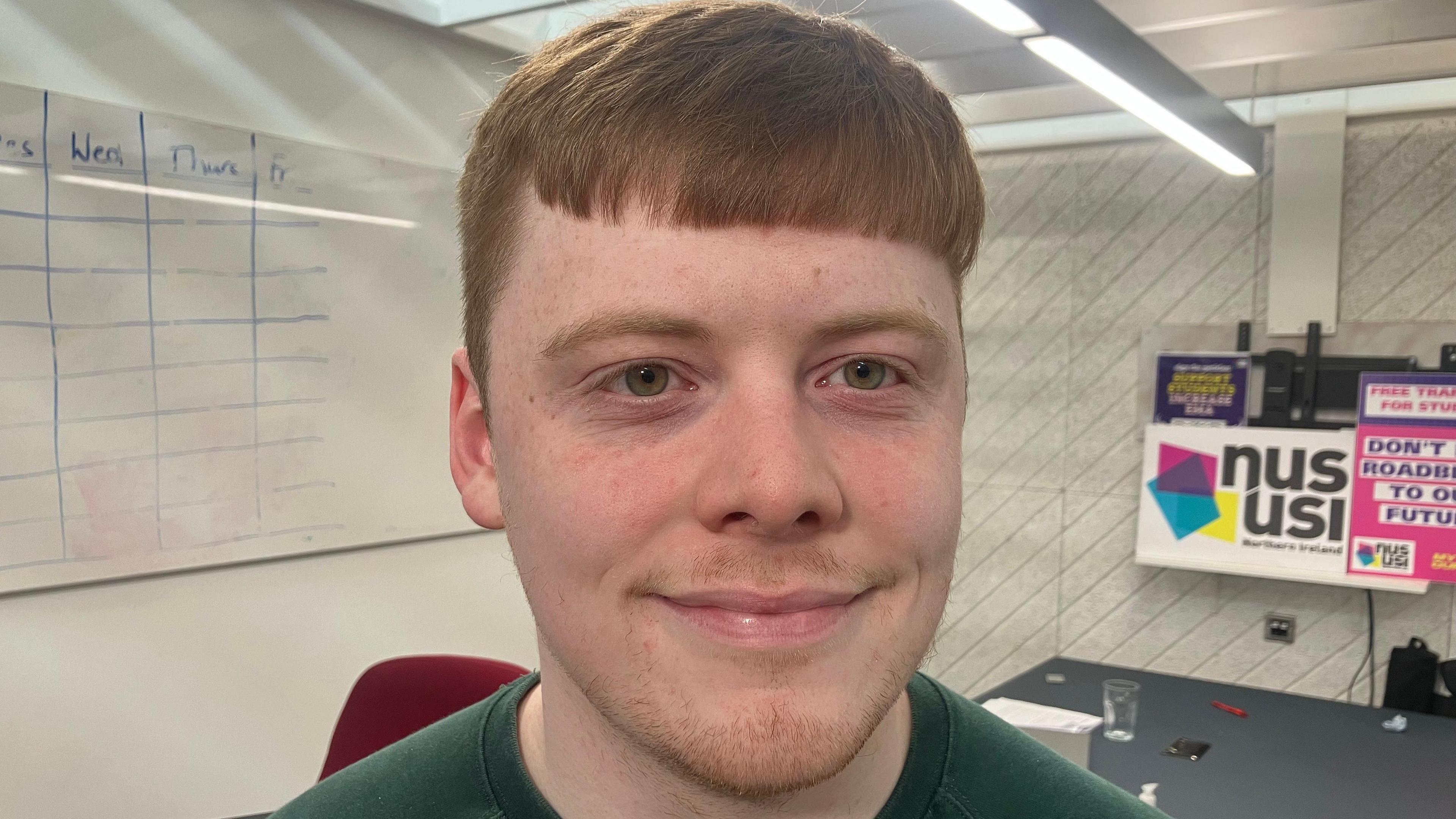 A man with ginger hair wearing a green t-shirt looks at the camera. There are NUS signs and a whiteboard behind him.