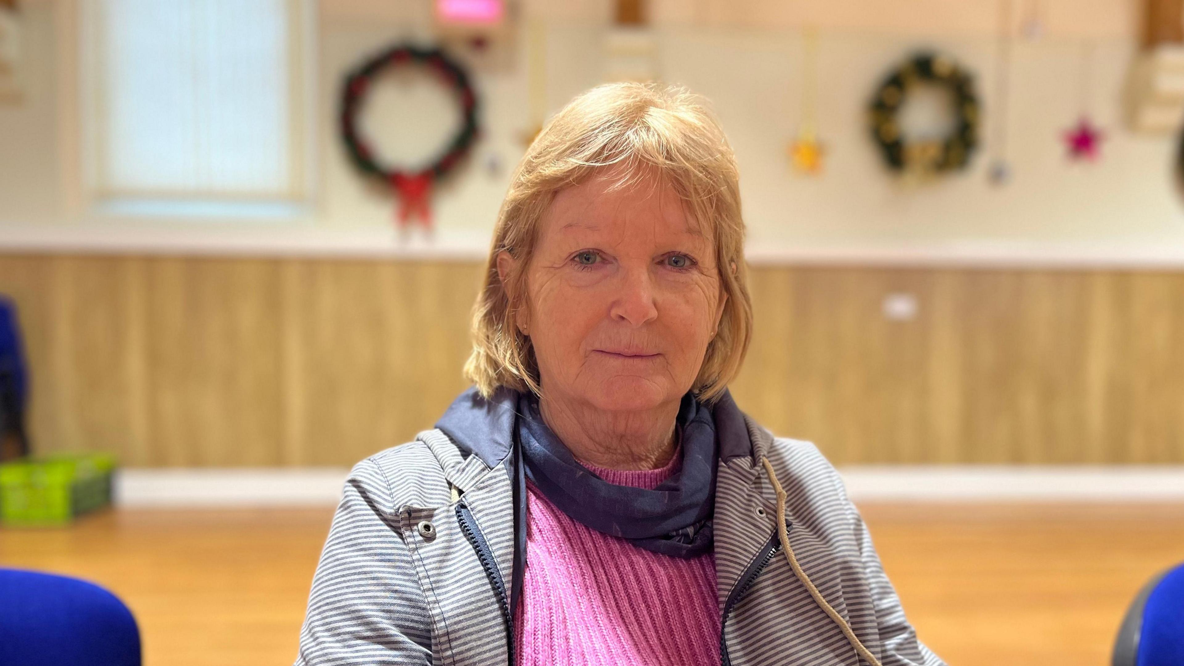 A blonde-haired woman sitting within a hall, wearing a pink jumper and grey open jacket. In the background are Christmas decorations.