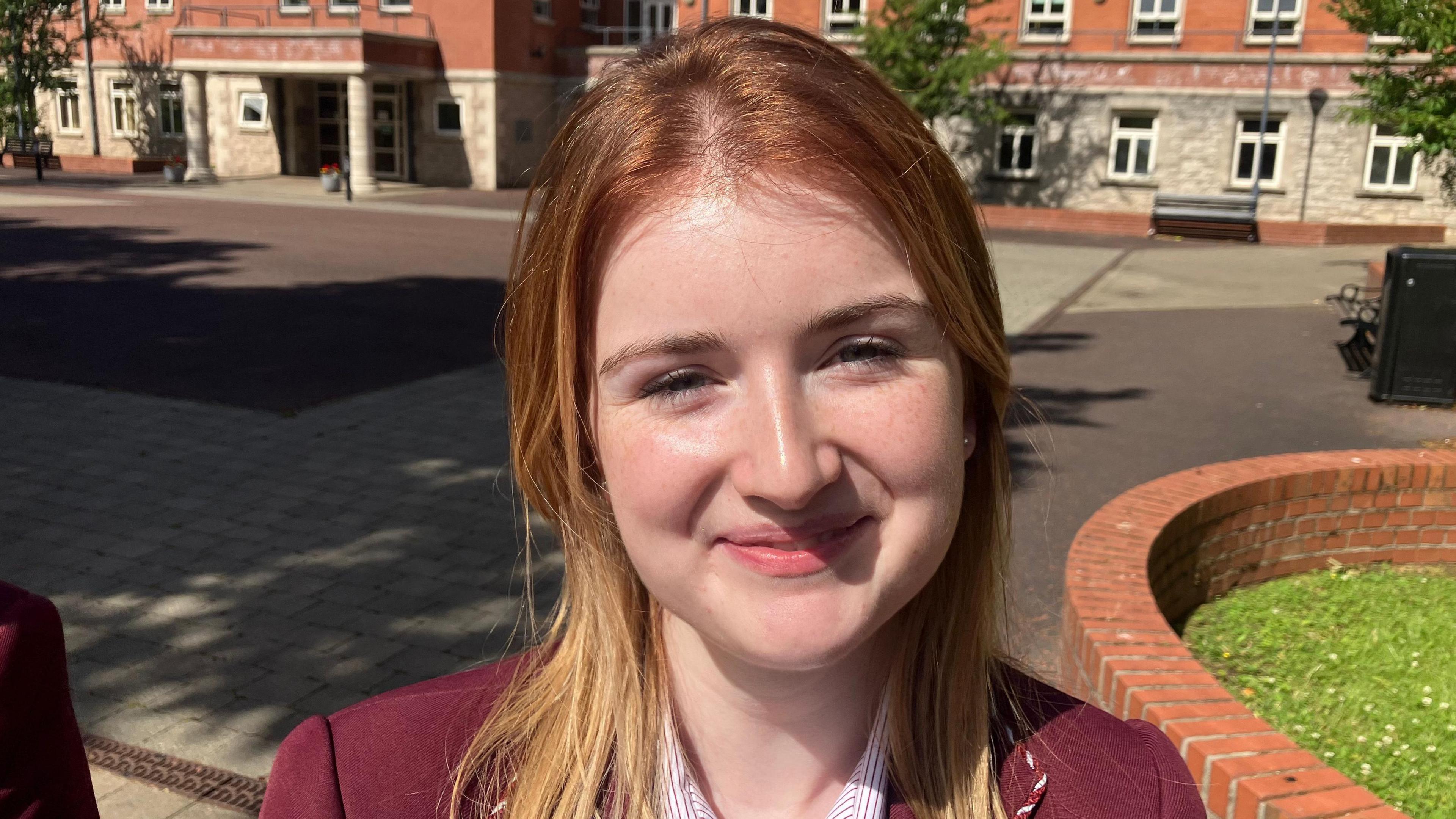 A young girl is smiling toward the camera. She has long blond hair. She is wearing a maroon coloured school blazer and a red and white stripe shirt. Behind her is an open stone courtyard and a number of red brick buildings.