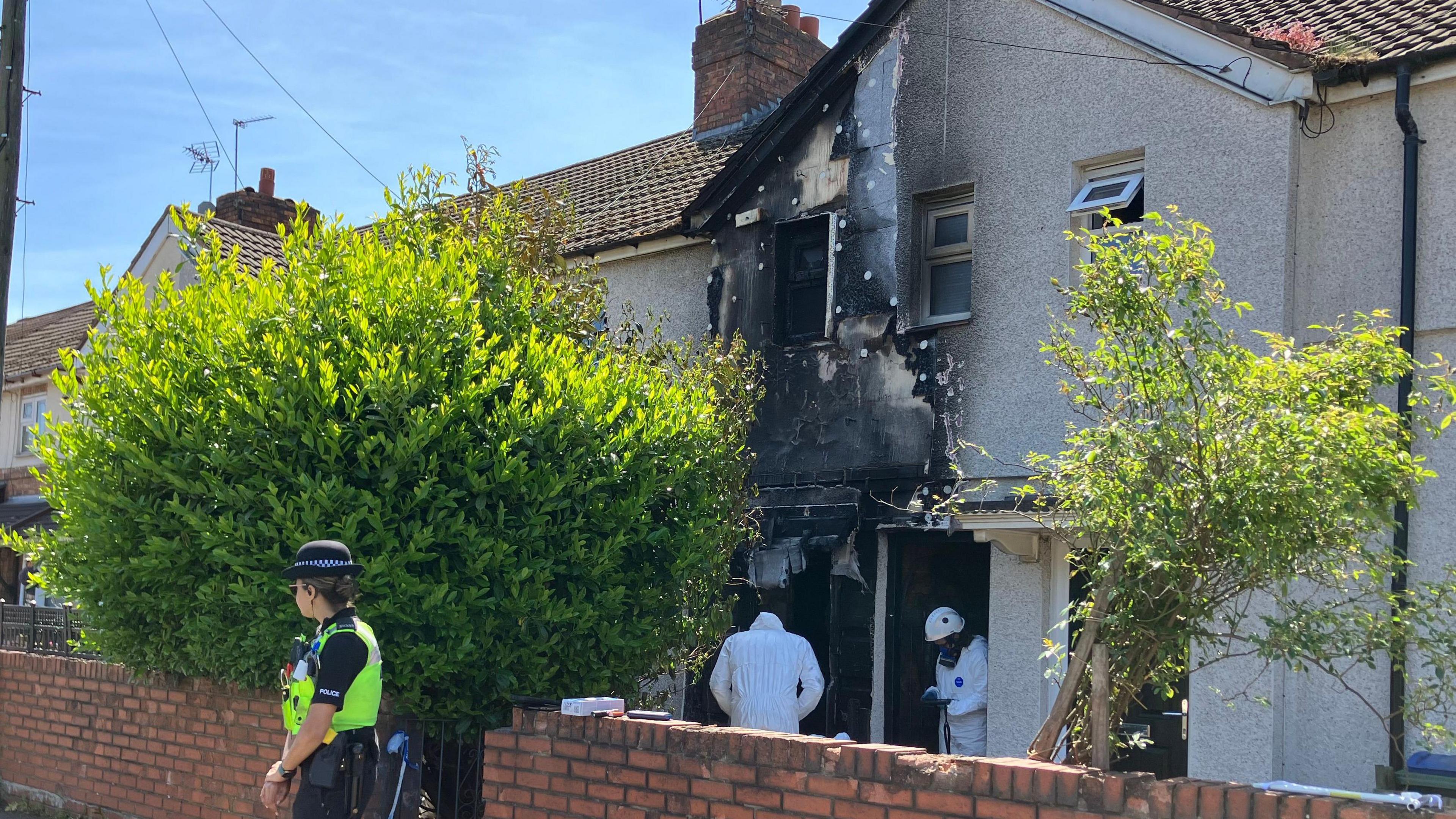 A terraced two-storey house shows fire damage on the groundfloor, first floor outer wall, window, part of the roof. Two people in a forensic outfit are at the front door whole a police woman is guarding the property from outside a waist-high brick wall.