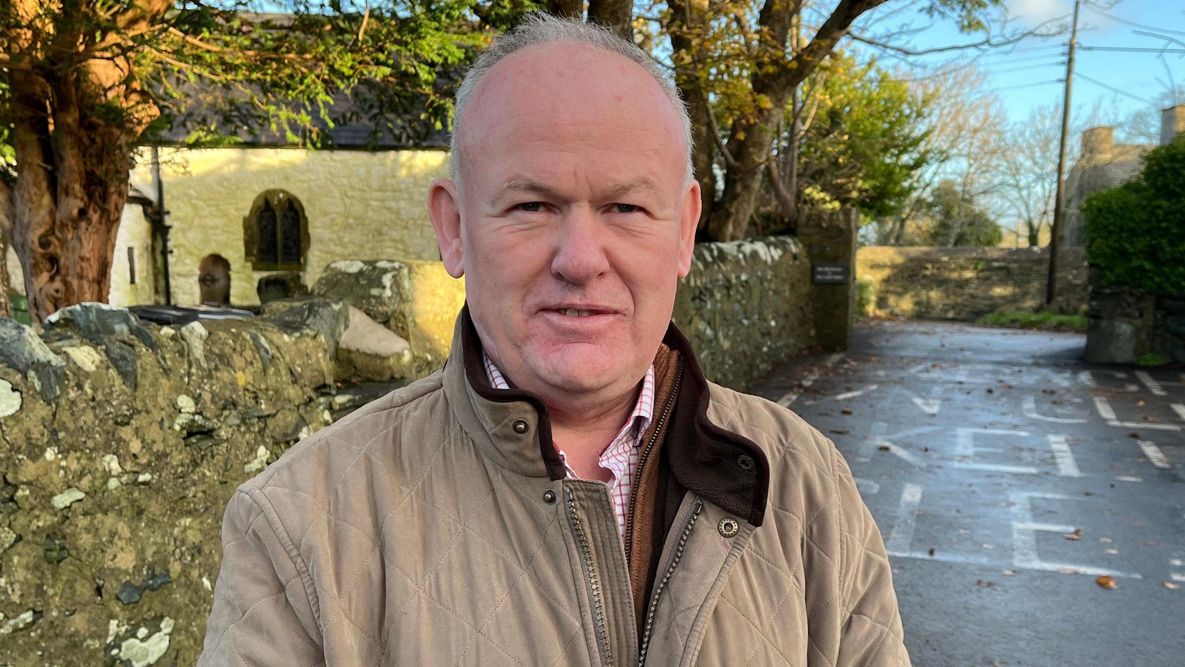 Llun o Aled yn sefyll ar stryd yn edrych ar y camera. Mae ganddo wallt llwyd a llygaid du. Mae'n gwisgo cot melyn