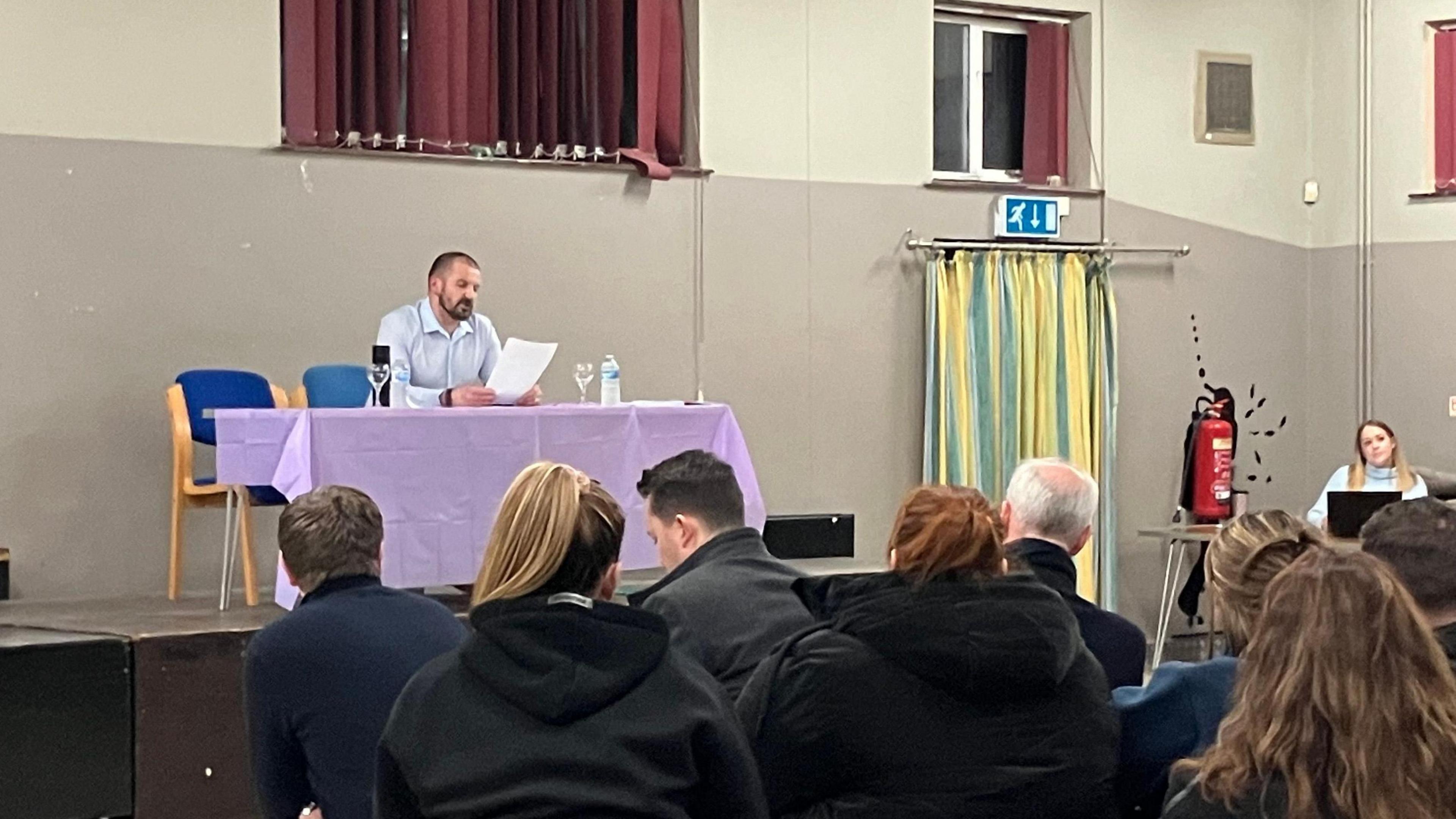 Roy Nineham sits behind a desk draped in a purple tablecloth. He is looking down at some papers he is holding and appears to be reading from. A number of people are sitting of chairs in front of him which face him. In the right corner is a woman behind a laptop, who is also facing the parents. 