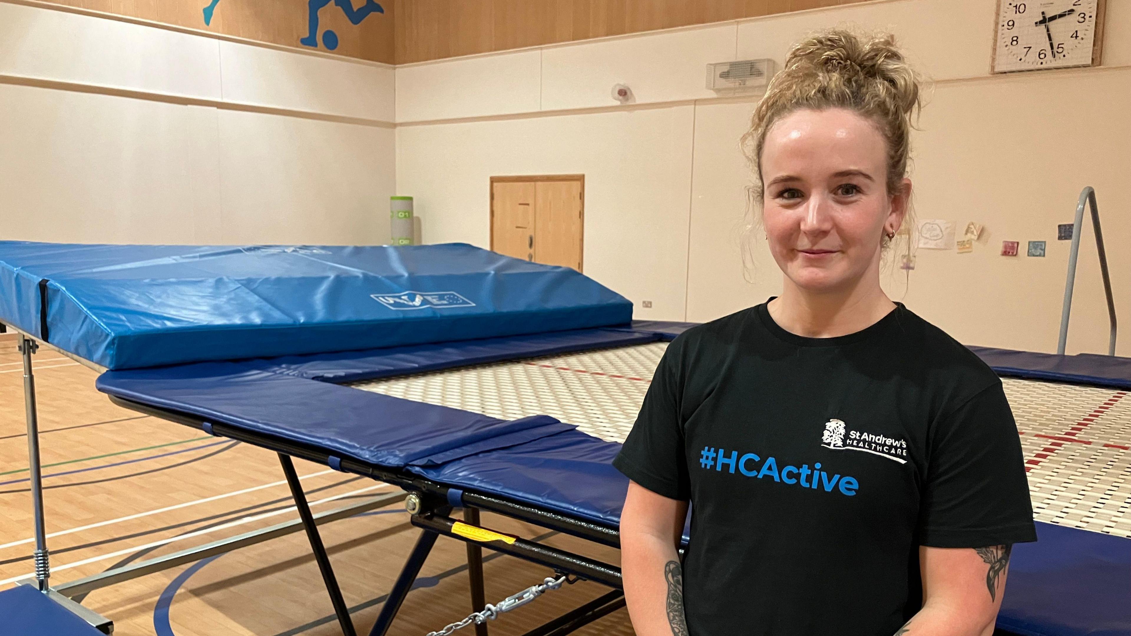 A woman with tied up blonde hair wearing a black t-shirt stands next to a wide trampoline with blue trim in a sports hall. 