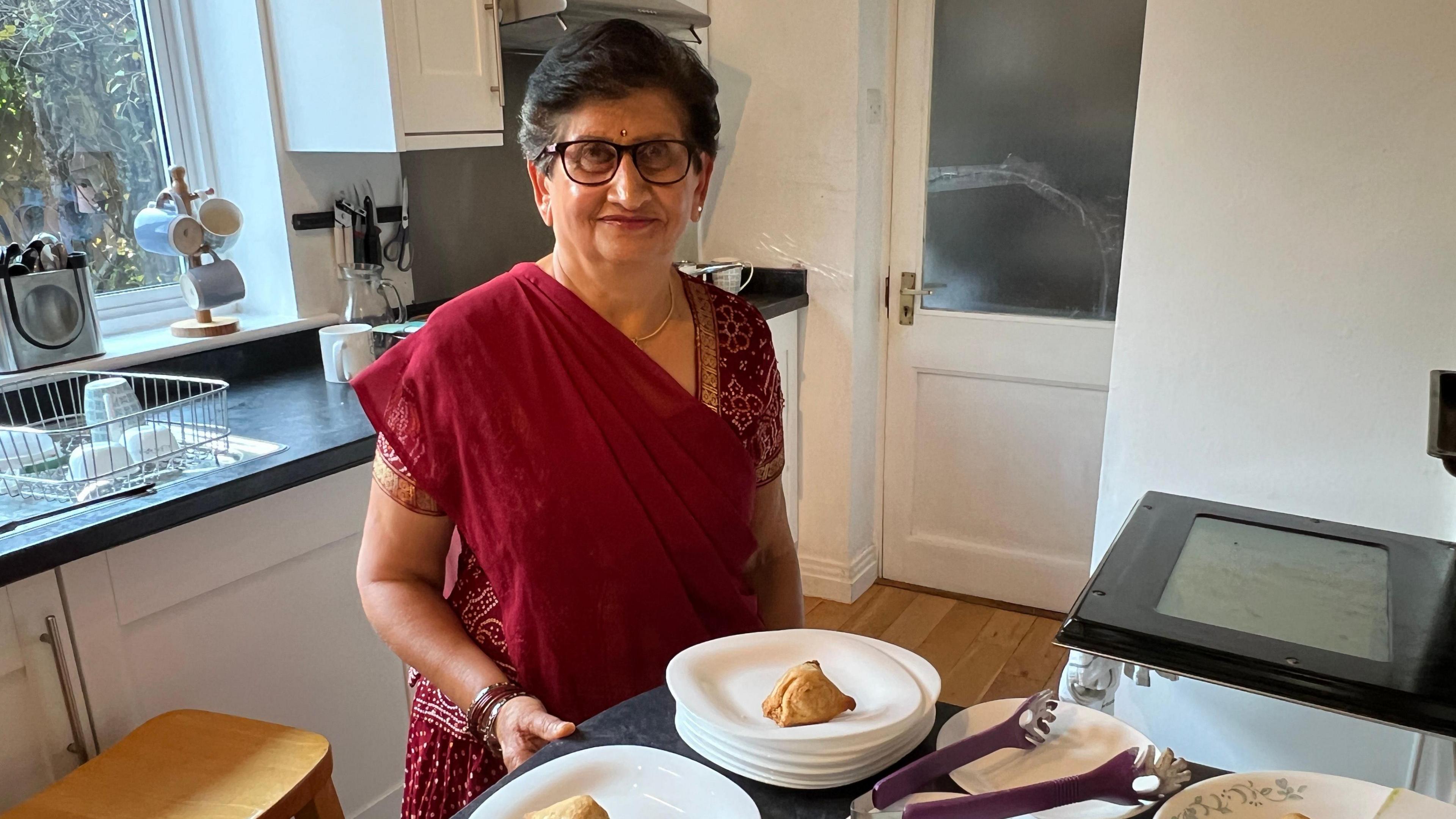 A woman in a red sari standing in a kitchen with samosas in front of her