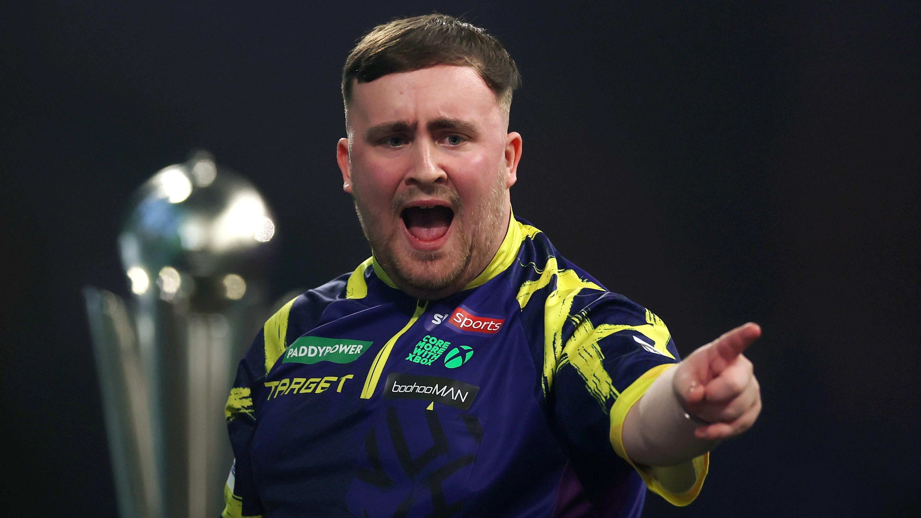 Luke Littler points with his left hand as he stands in front of the World Championship trophy