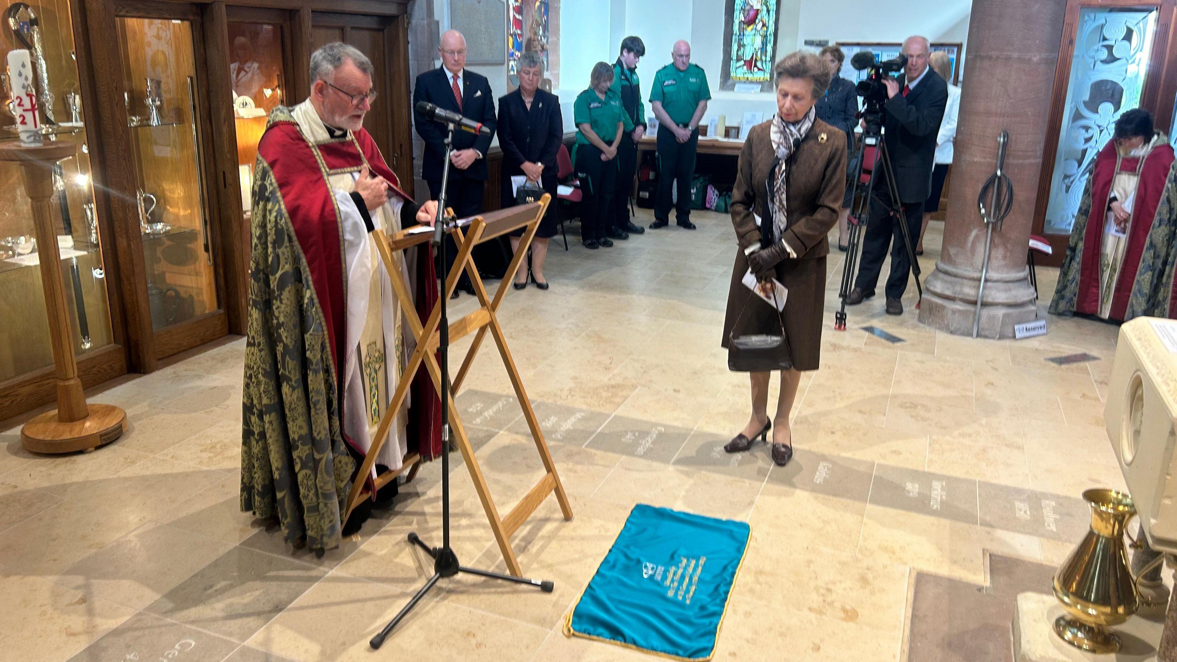 Princess Anne looks down at the floor where there is a small blue cloth, which the Dean of the Cathedral Nigel Godfrey is standing at a lectern in front of it speaking.