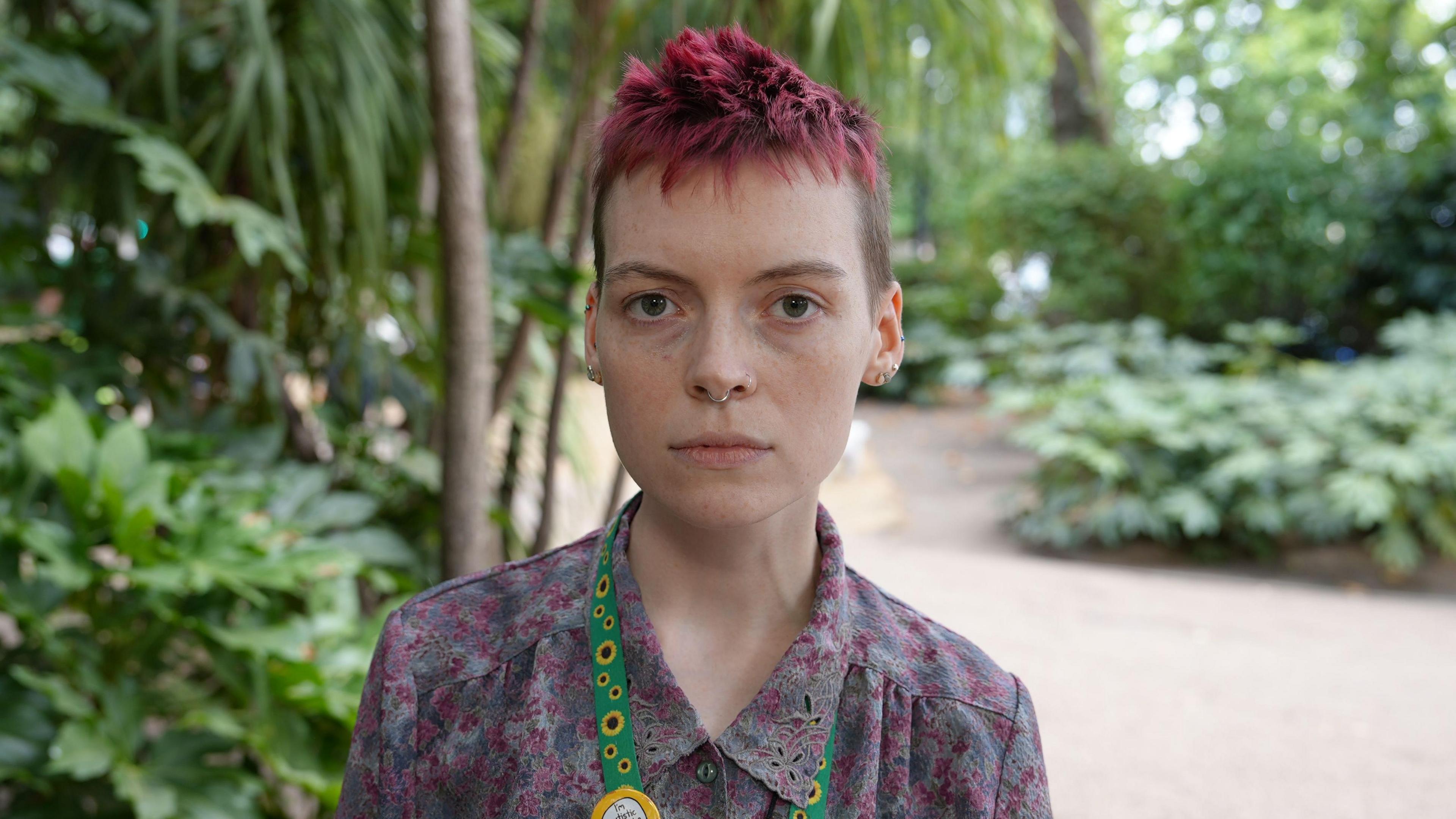 A person with short spiky hair looking at the camera. They are wearing a floral shirt and a green lanyard with sunflowers on it. Behind them is a path and greenery.
