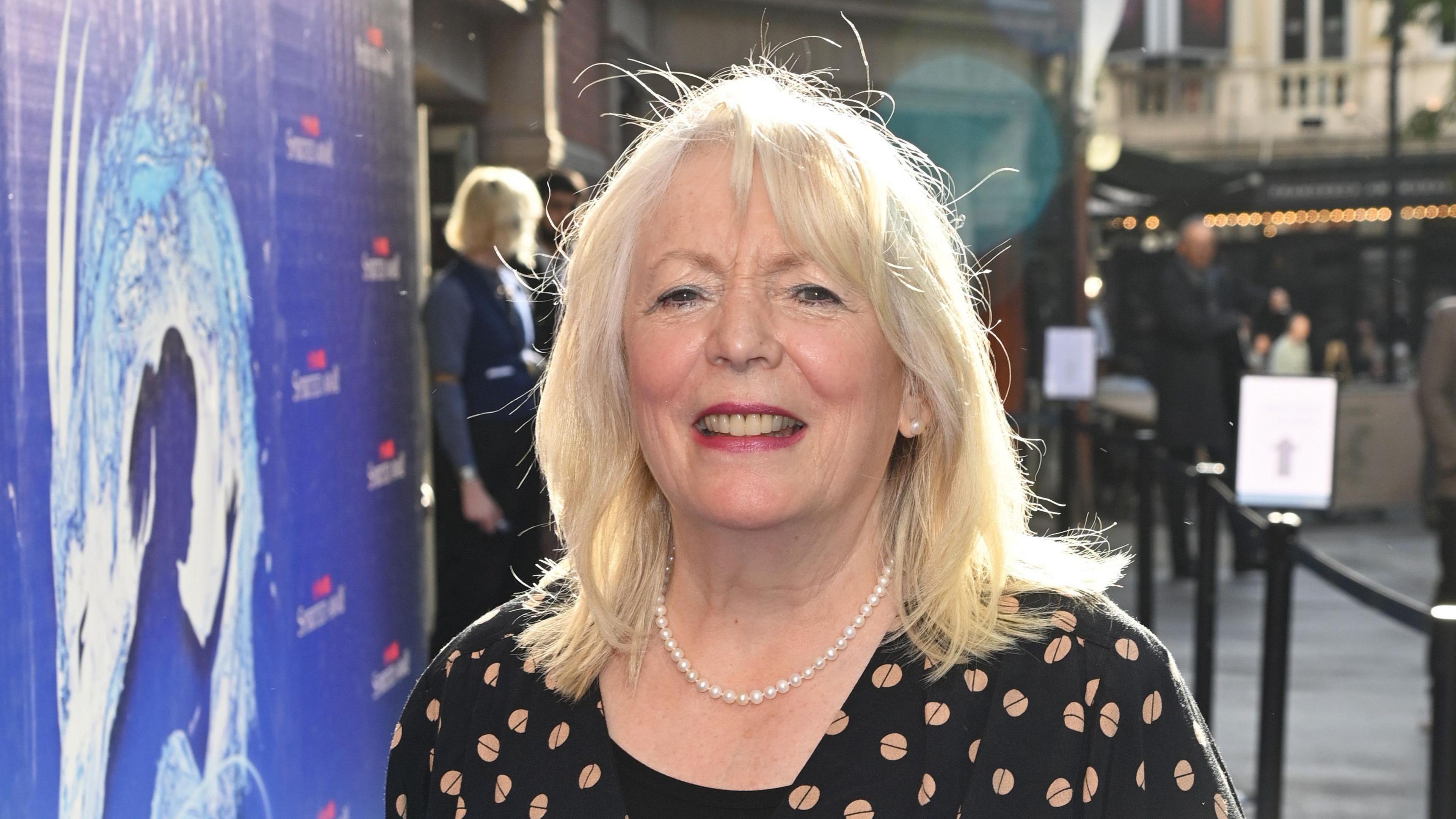  Alison Steadman smiling and wearing pearl beads and black dress with peach dots. She is stood on a red carpet at a premiere 