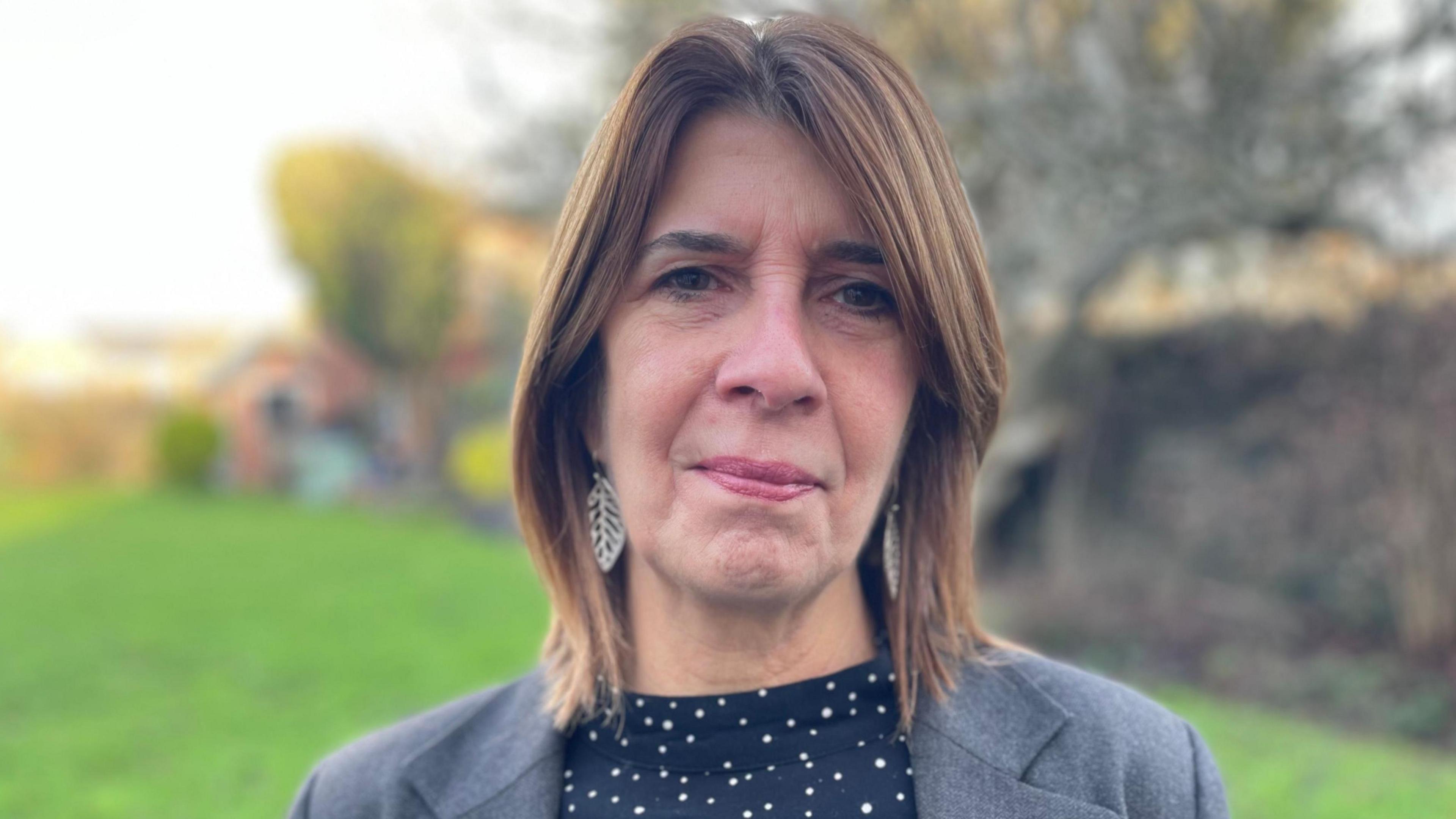 Rosie Lewis o Unsain gyda gwallt brown yn gwisgo blows ddu gyda sbotiau gwyn, siaced lwyd a chlustdlysau arian, siap dail.