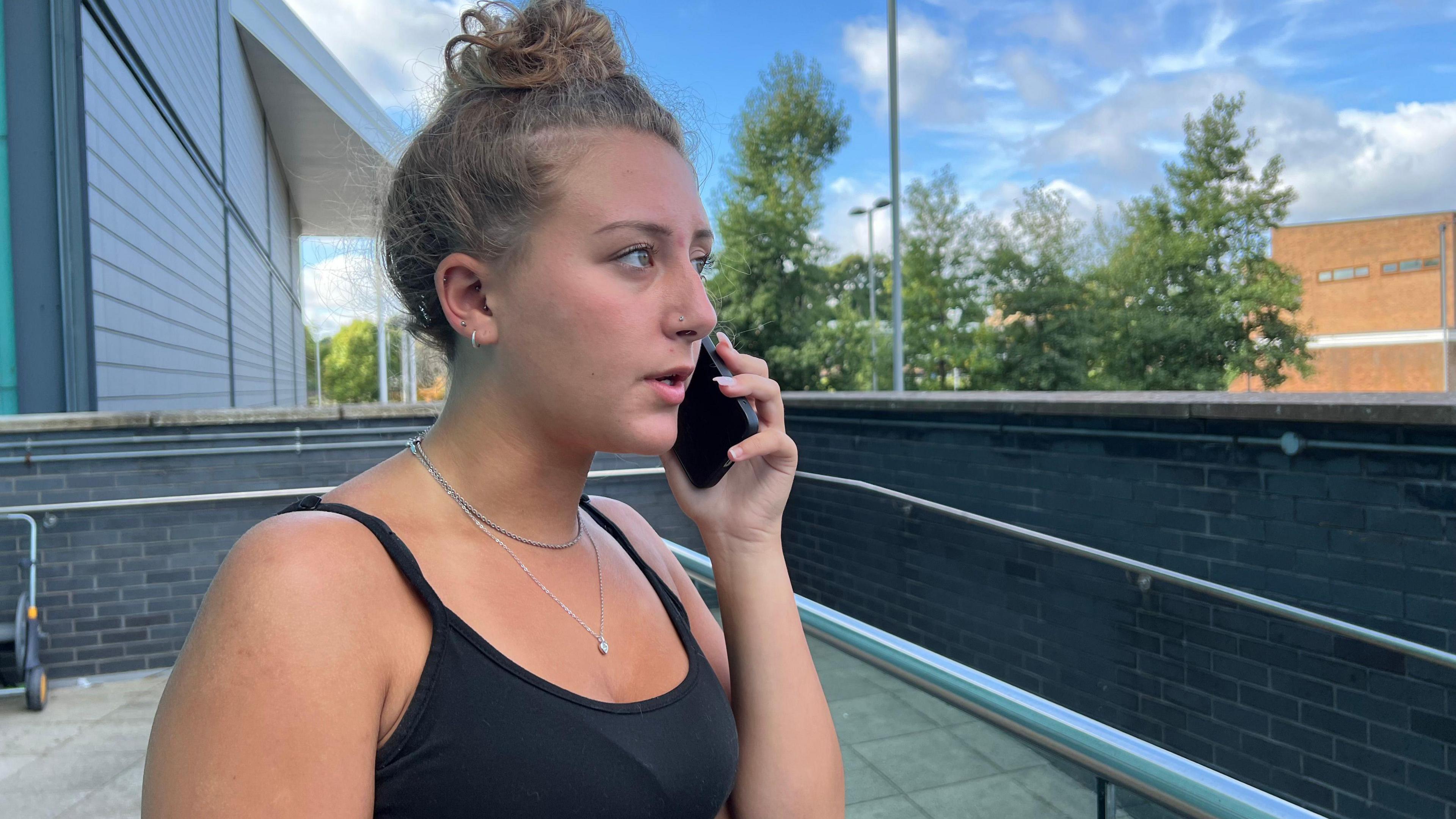 Caitlyn Wyeth is standing outside on a balcony, speaking on the phone. She has light brown curly hair which is tied up into a bun on the top of her head. Caitlyn wears a black camisole top with two silver necklaces.