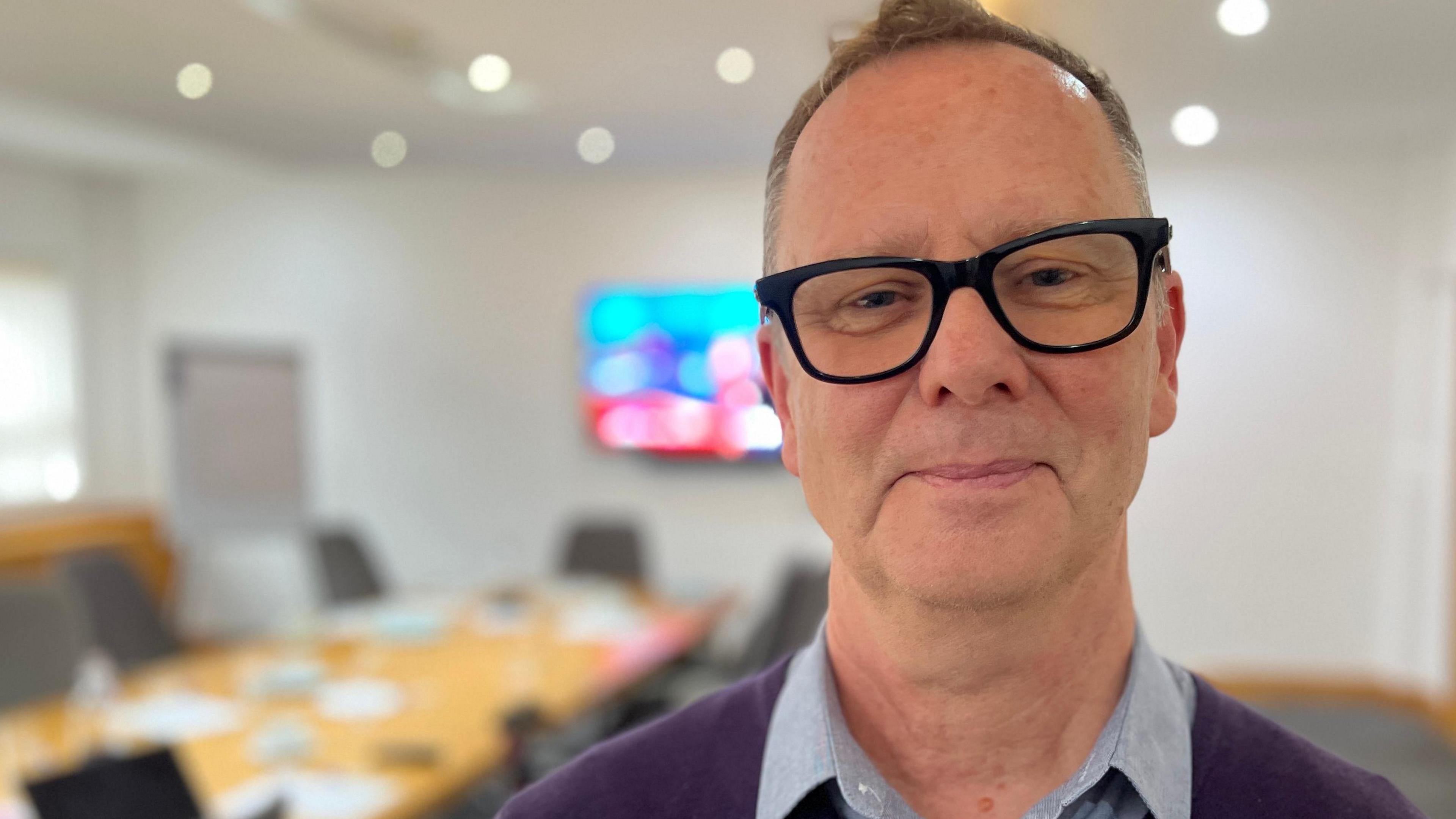 A smiling Neil Woodbridge looks directly at the camera. He is wearing large glasses, and a purple jumper with a blue shirt. In the background is a TV and empty chair around a boardroom table.