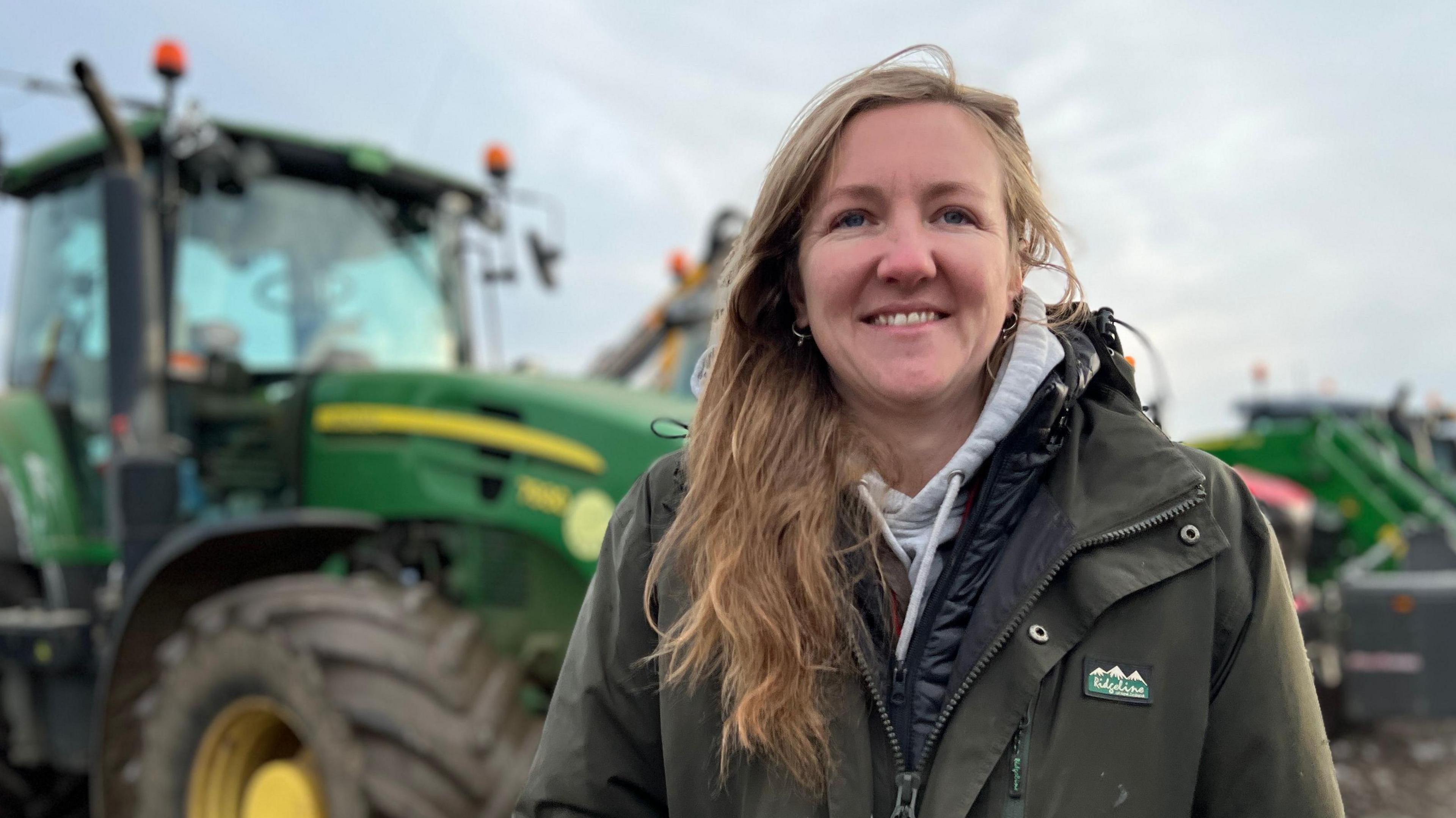 Kate Bowsher is stood wearing a green coat with a grey hooded jumper underneath. She is standing in of a row of tractors with a large green tractor clearly visible in the background over her right shoulder. 