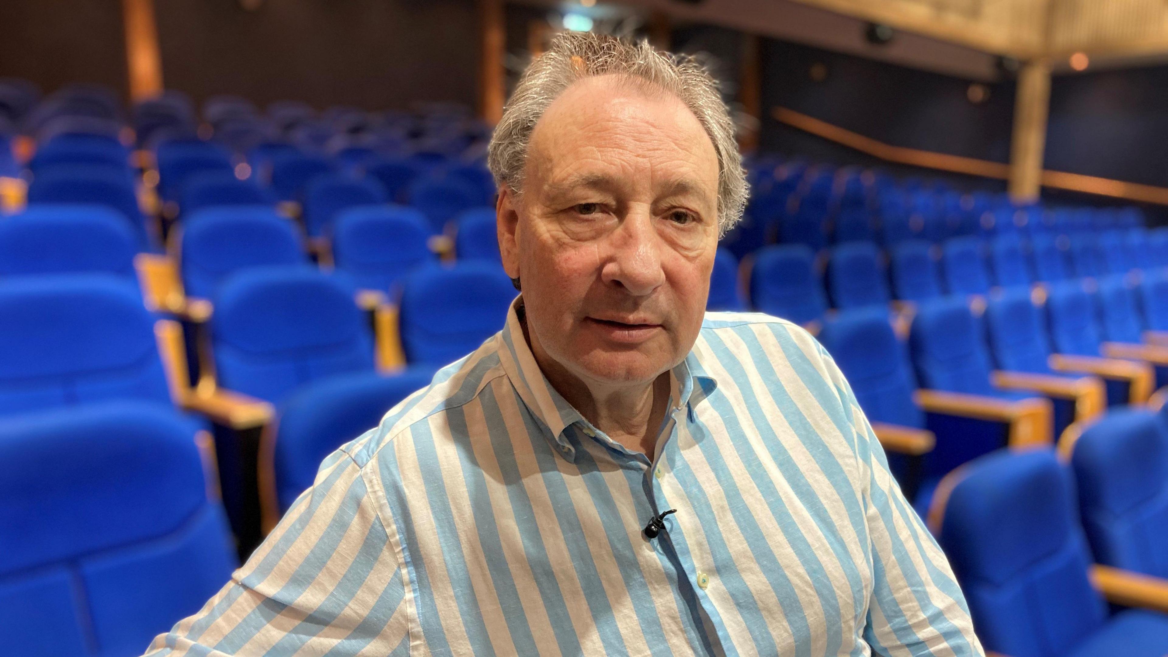 A man with grey hair and wearing a stripy blue and white shirt sitting in a theatre, with rows of blue audience chairs in soft focus behind him.