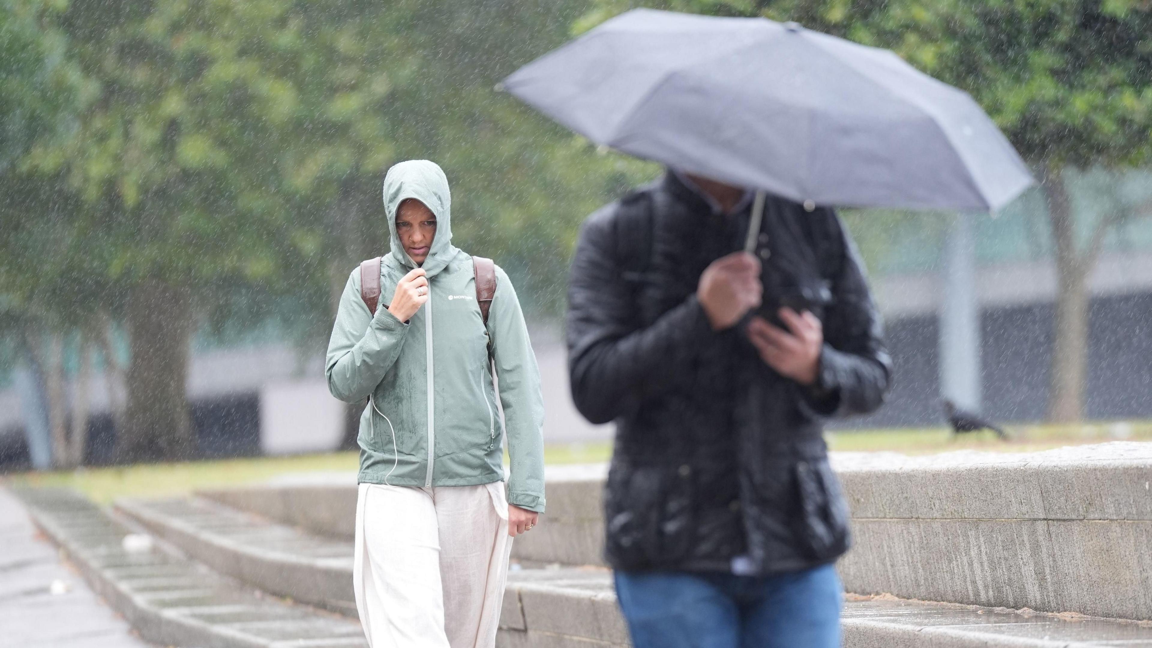 Two people walking through a park, with trees. in the background. One is wearing light clothing with a hood up. the other is wearing dark clothing, with an umbrella. It is raining.