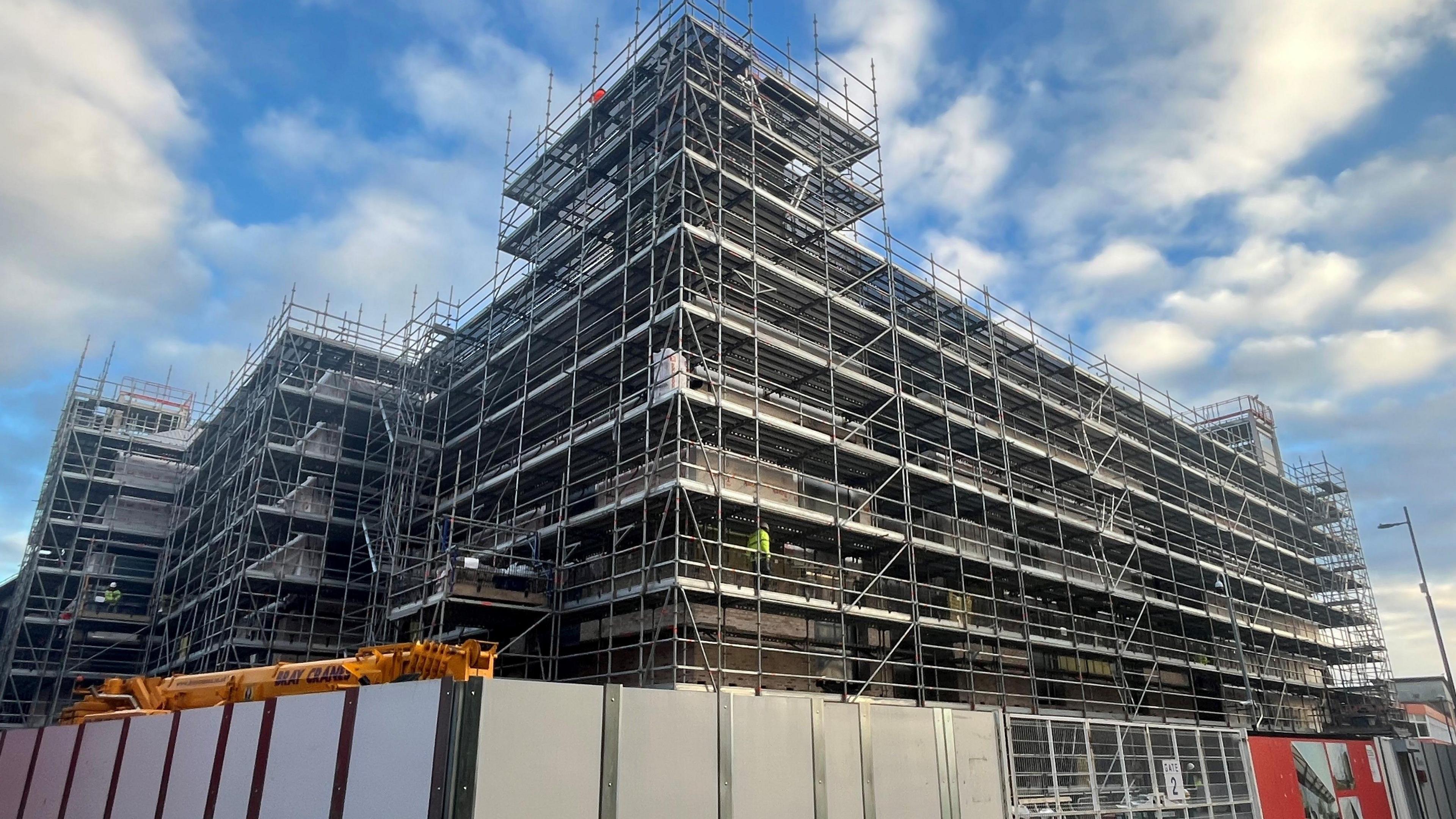 The building site of the multi-storey Culture House, covered in scaffolding. Two workers in helmets and high visibility clothing are walking on the structure. 