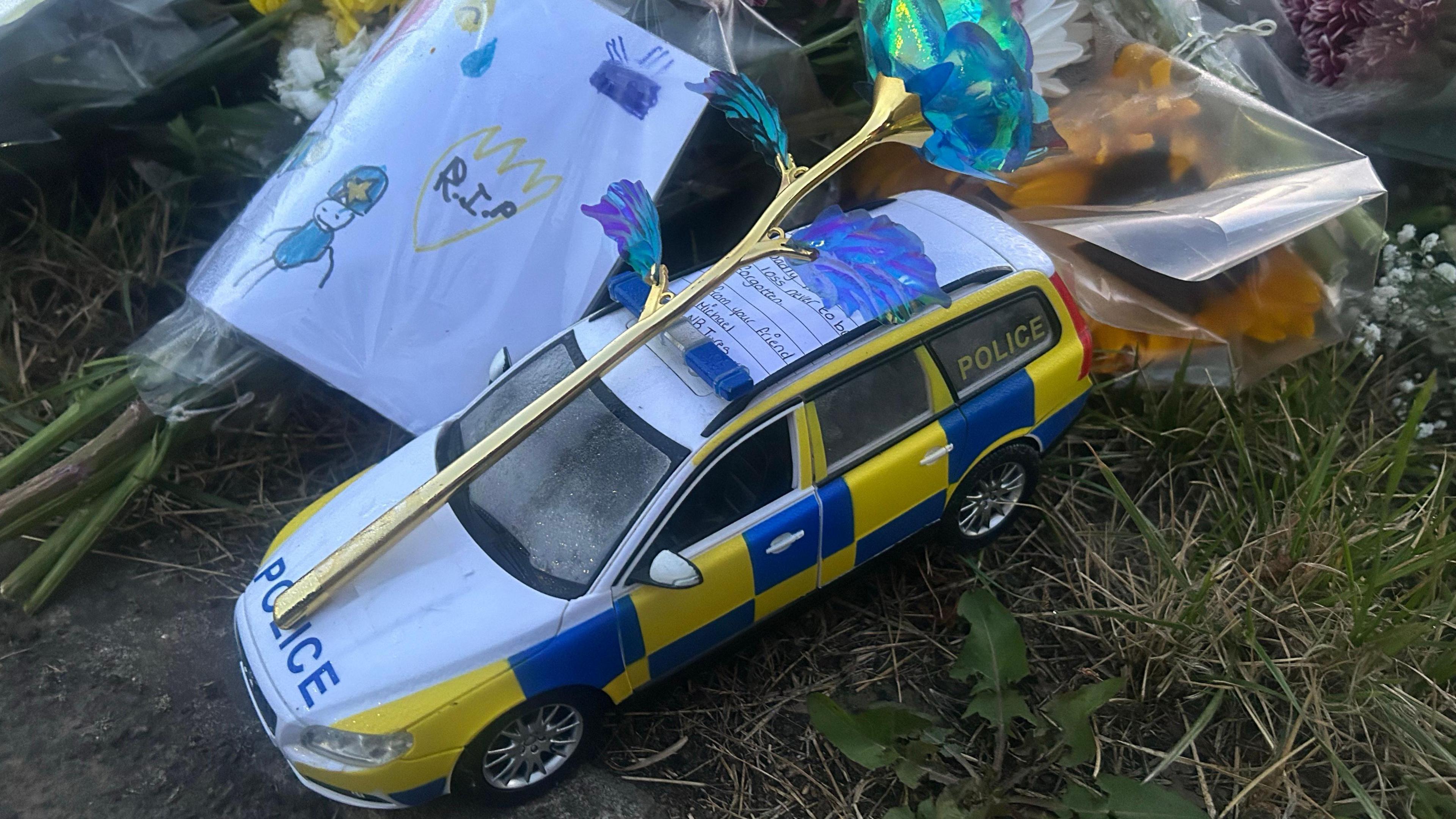 A yellow, blue and white toy police car is set among the floral tributes. There is a plastic flower set on top of it as well as a handwritten note. There is a drawing of police officers and the word "RIP" on a piece of paper wrapped around another bouquet of flowers on the left.