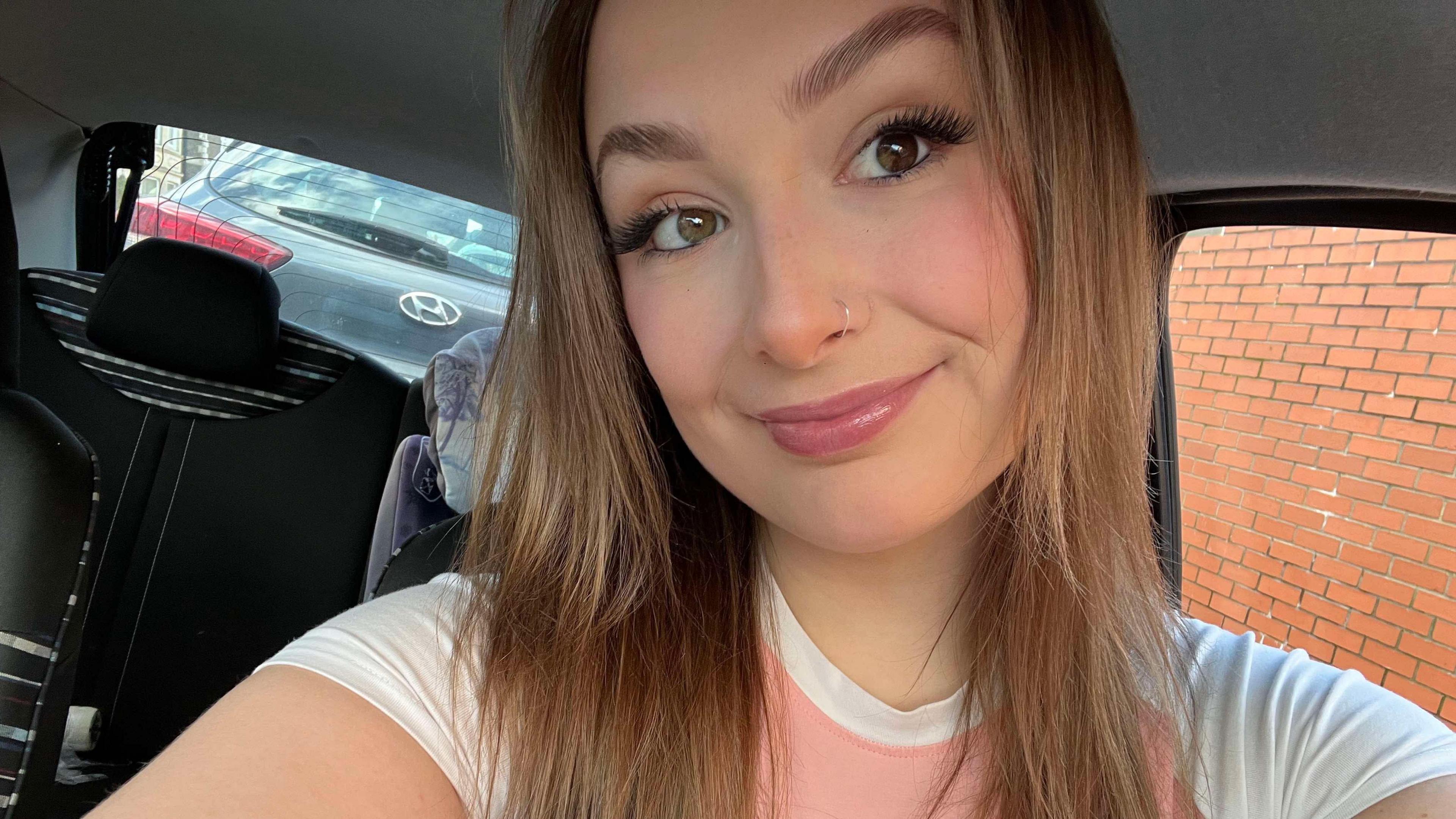 Layla Cahill Harris smiles into the camera. She is wearing a pink and white t-shirt and is in the car. She has long brown hair which frames her face, pink lipgloss on and a nosering. 