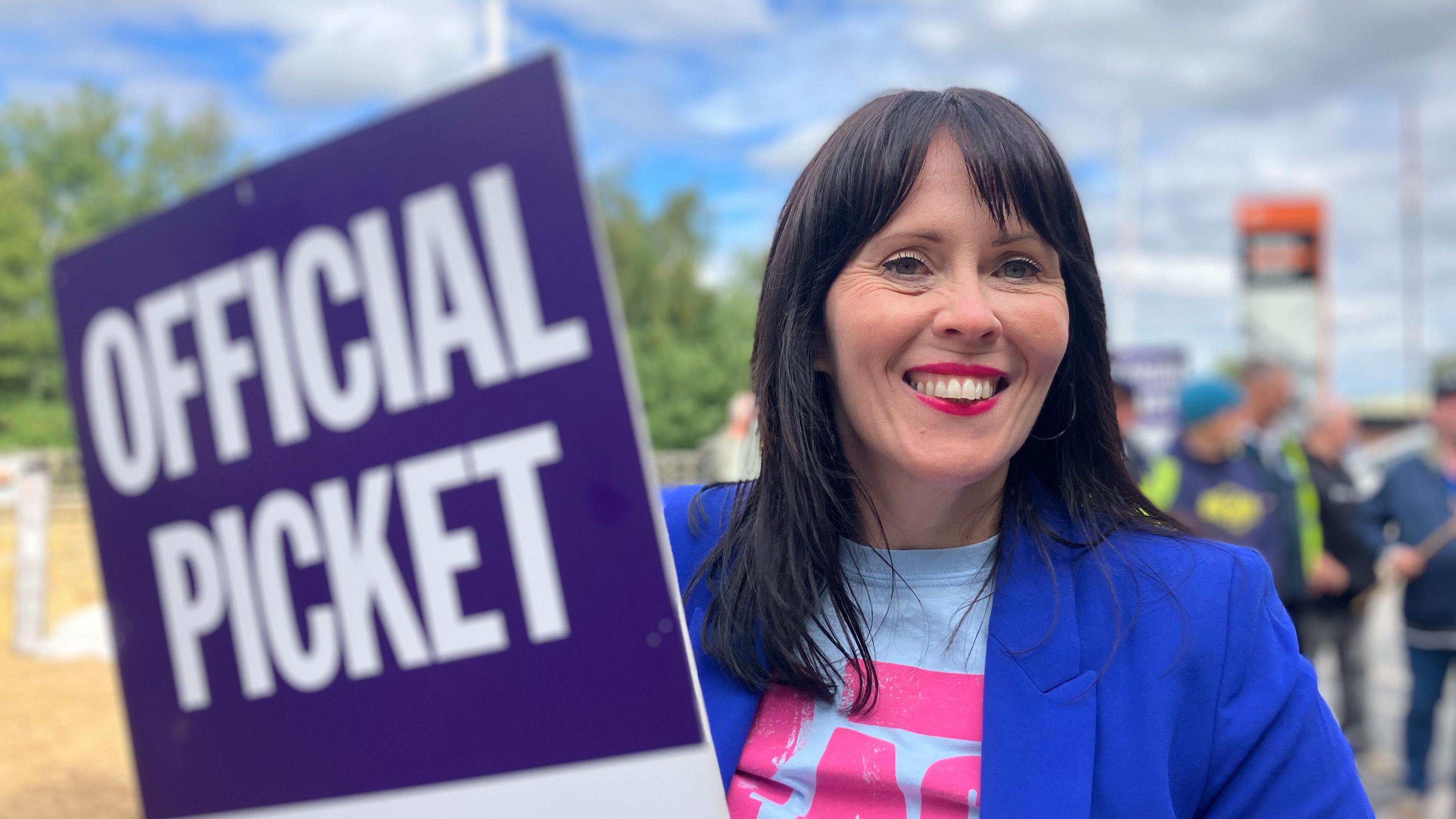A headshot of a woman, smiling at the camera, with red lipstick, black hair, and a bright royal blue blazer. She is holding a purple sign which says "official picket" in white capital letters.