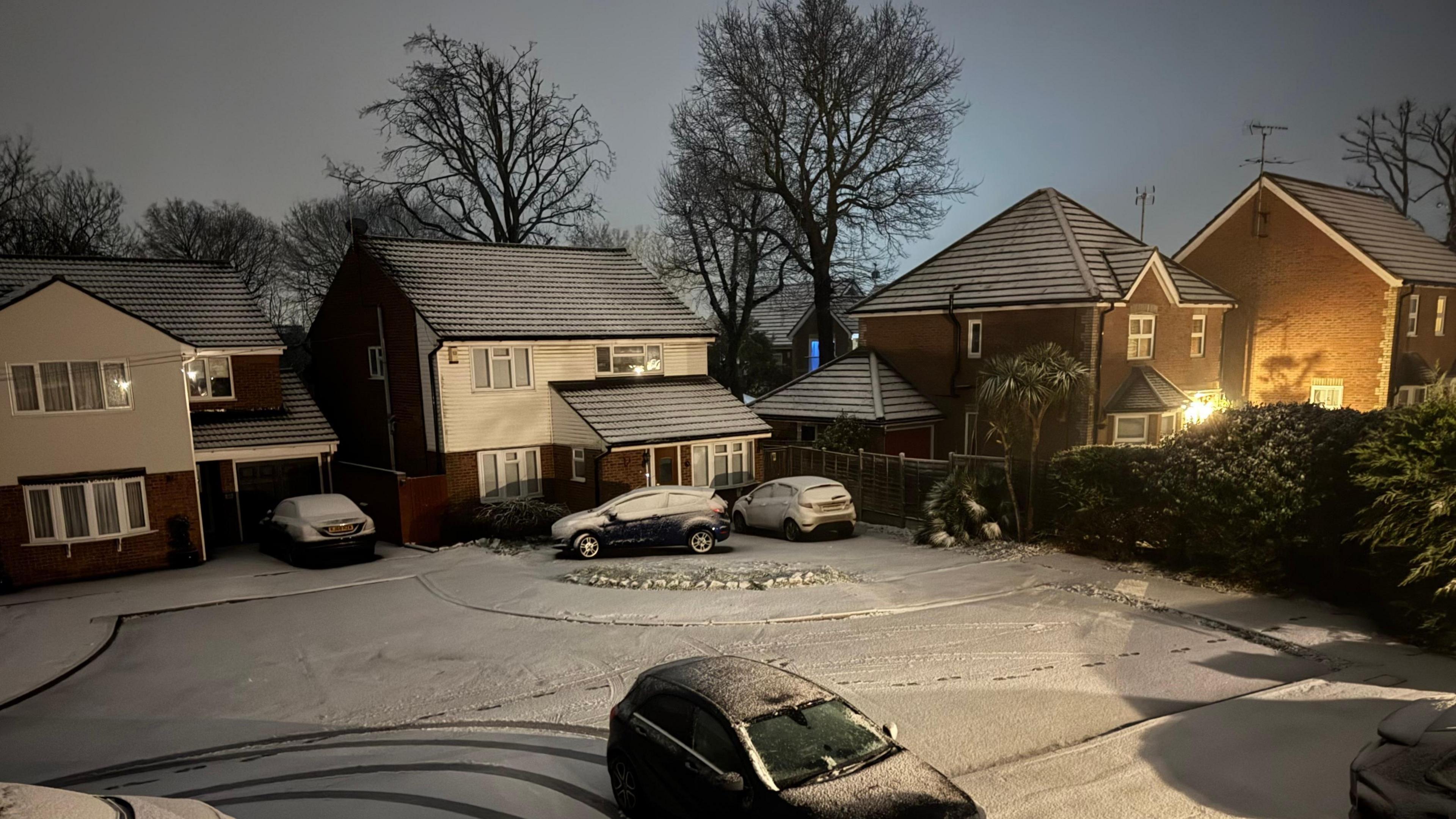 A road covered in snow, the snow is on cars, houses, trees, and with some lights on homes. There are tyre tracks on the road. 