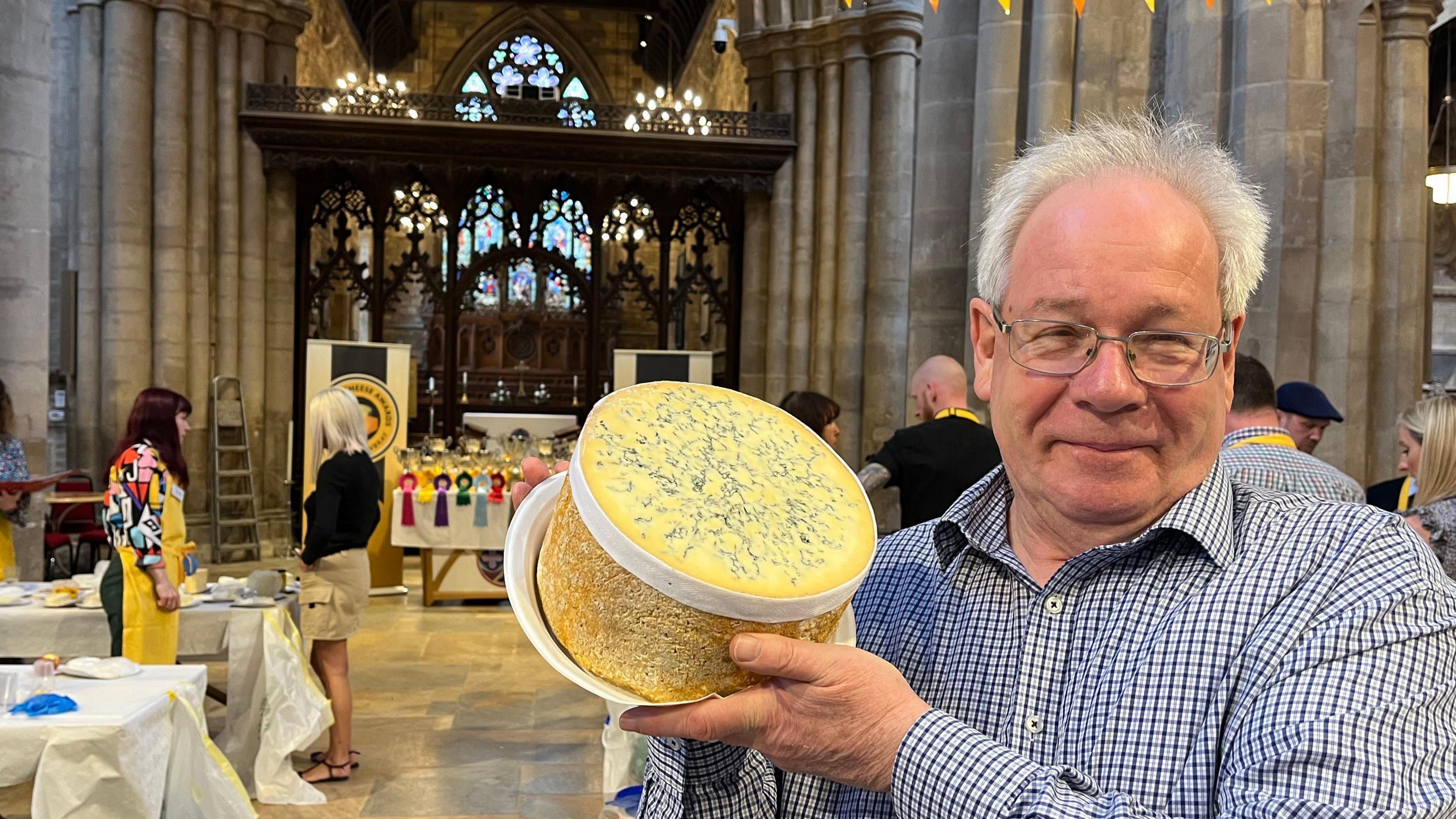 Man in glasses with checked shirt holding wheel of blue cheese