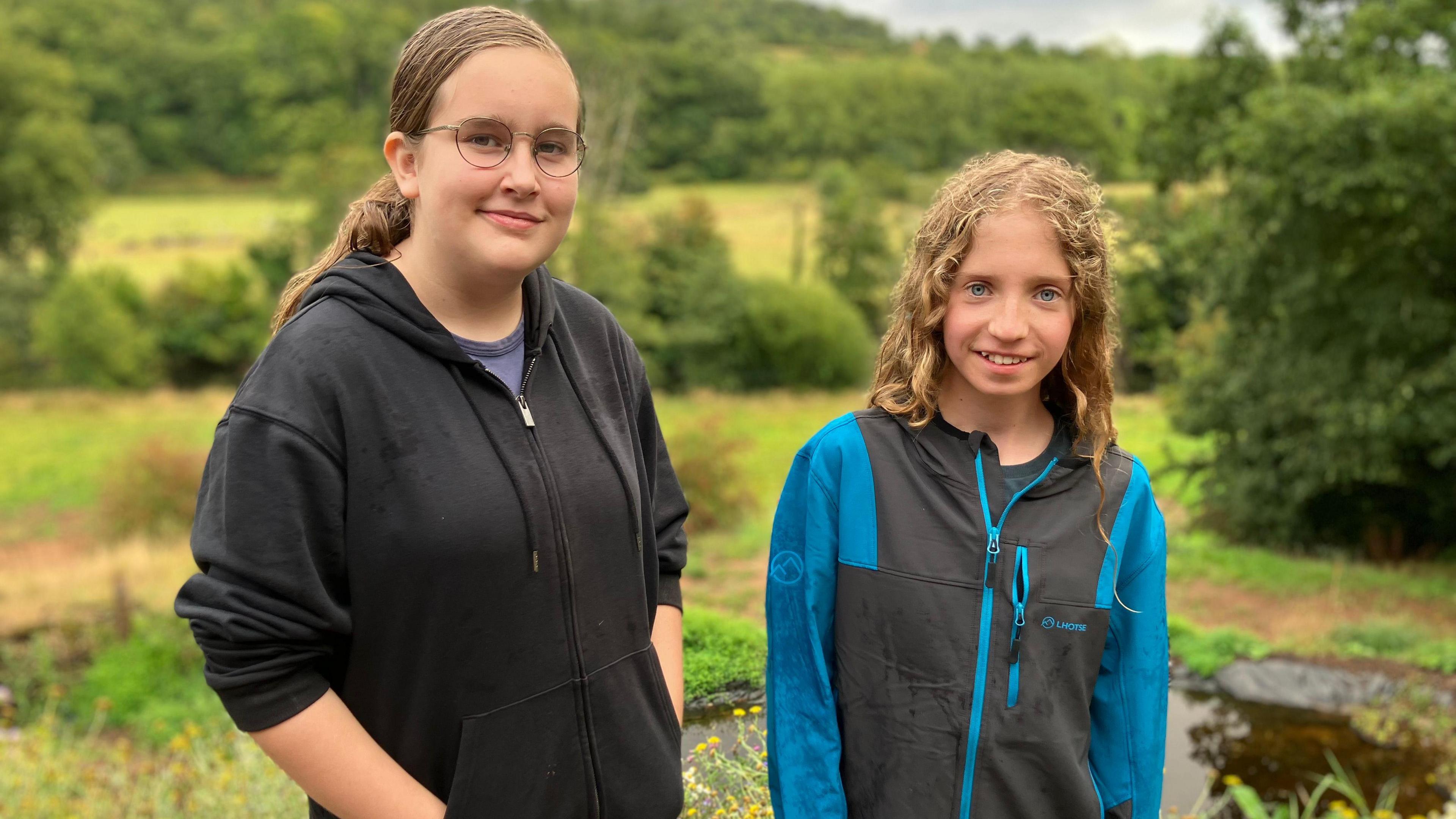 Emily, on the left, with her brown hair in a ponytail. She is wearing a black zip up hoodie and glasses. Eva, right, wearing a blue and grey zip up jacket. She has blonde curly hair which is down. Both are looking at the camera and smiling. In the background is green grass and bushes. 