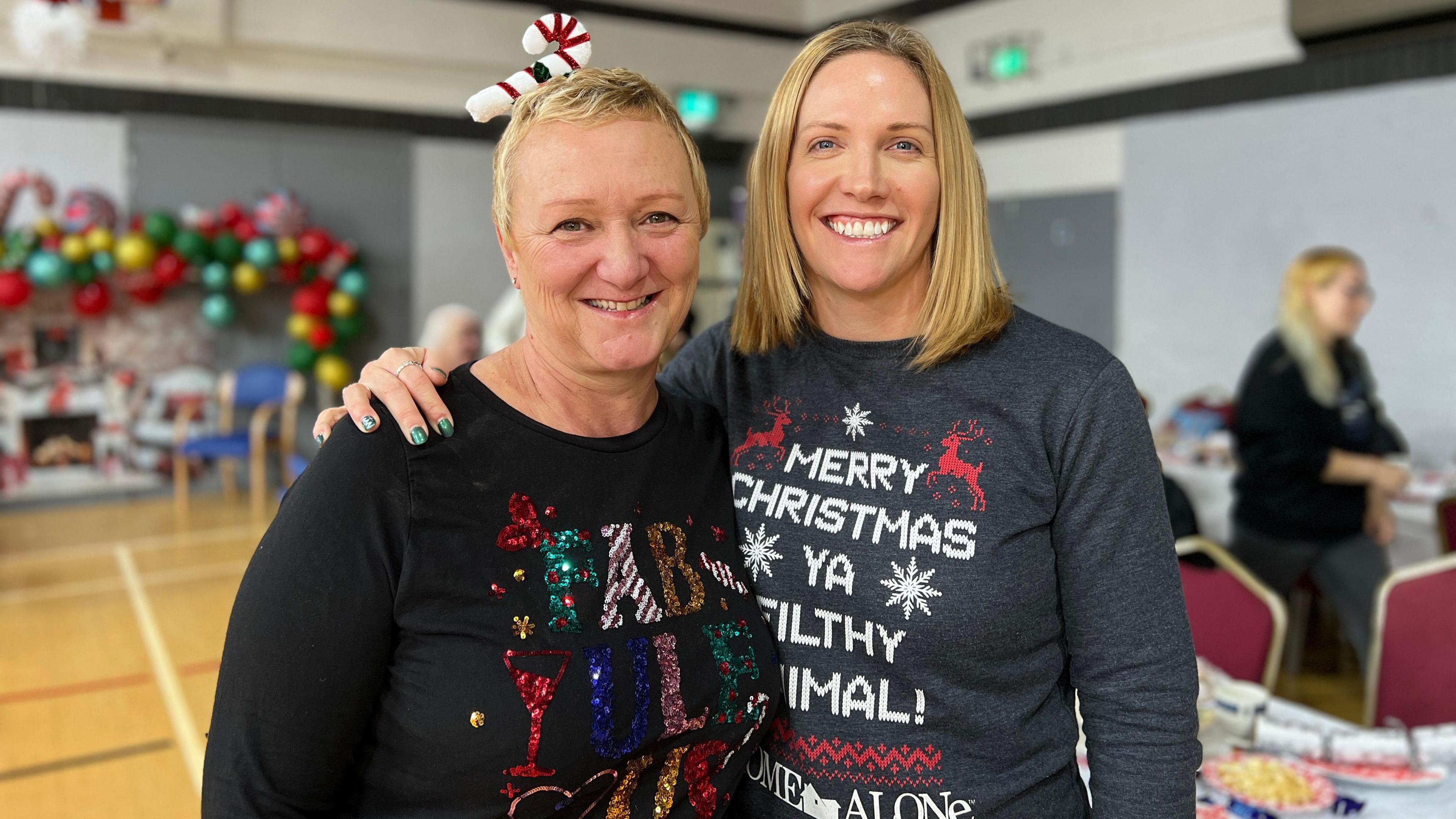 Jo and Jen smile at the camera and they're standing in a youth centre hall with dinner tables and chairs behind them. Jo is to the right and is slightly shorter with Jen putting her arm over her shoulder. They're both wearing Christmas jumpers.