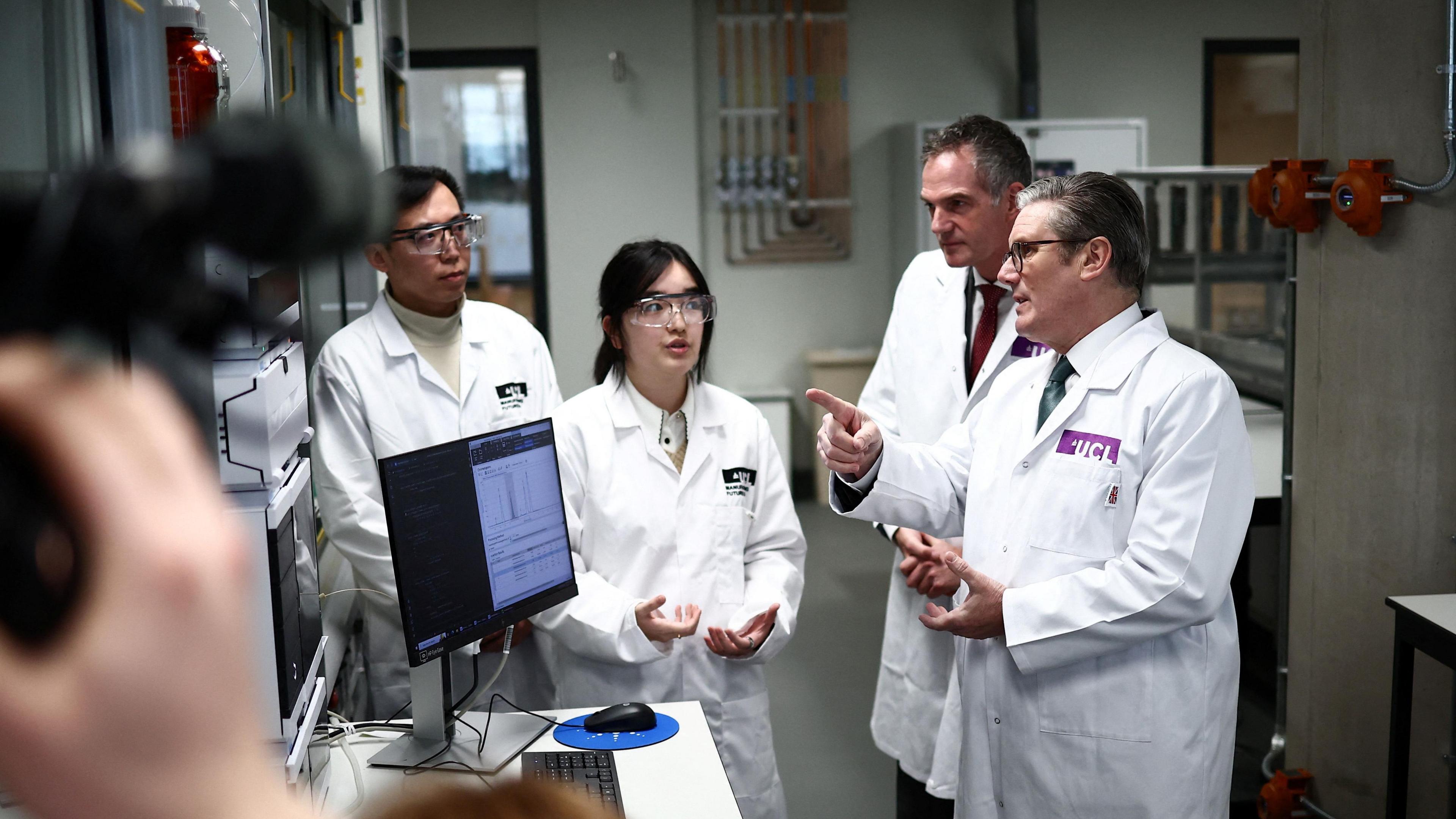 Four people wearing white lab coats mid-way through a discussion, all standing up in a laboratory-style setting. A computer screen is switched on at a desk in front of them. The man on the right of the image is Sir Keir Starmer. He is pointing towards something out of frame over to the left and appears to be speaking. The next person from left is the secretary of state for science, innovation and technology, who looks to be listening intently. Next along from are two unidentified people, one of whom appears to be about to speak. These other two people are both wearing safety goggles.