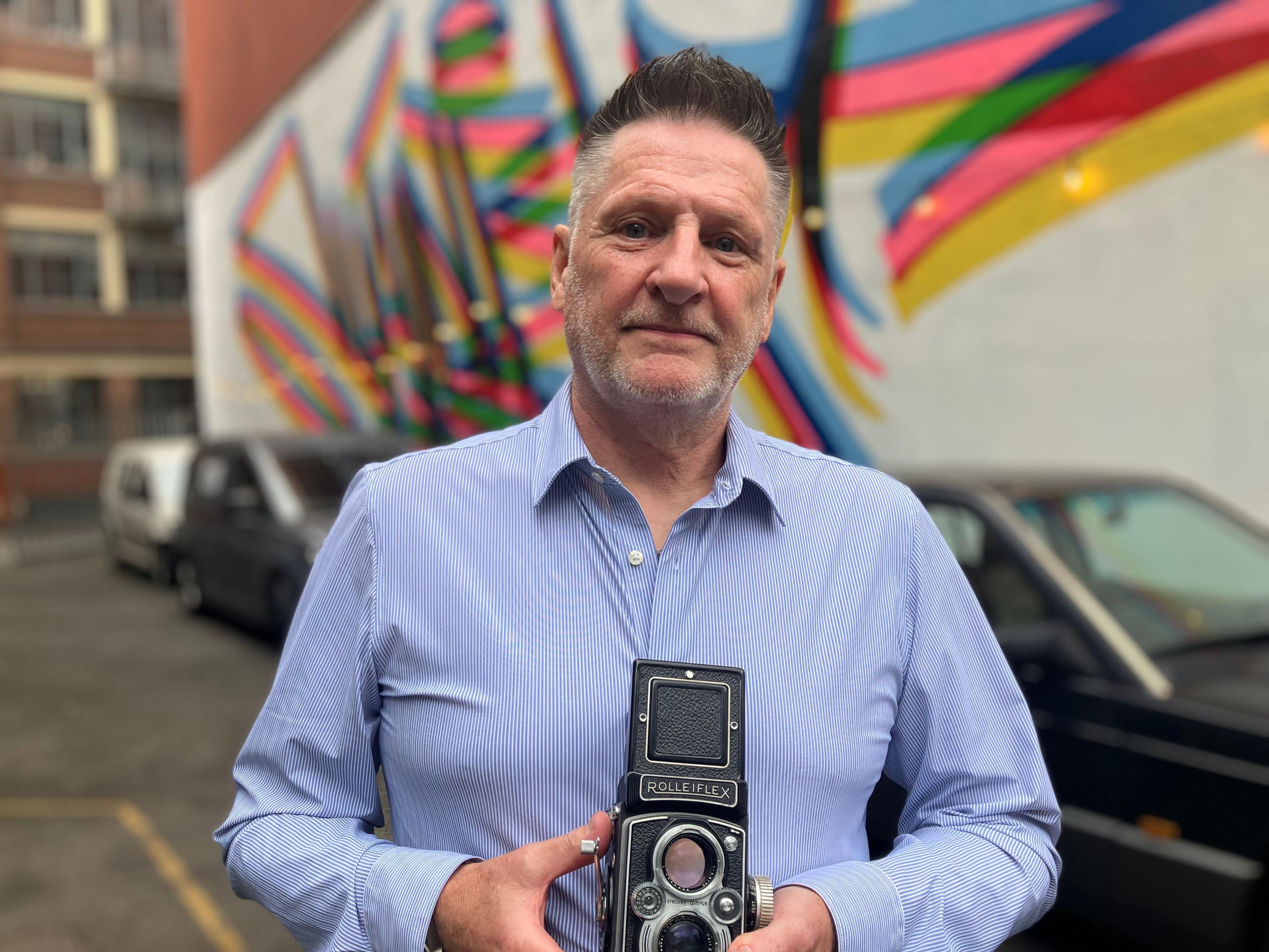 Wayne Large stands in front of a colourful mural. He is wearing a blue shirt and holding his vintage Rolleiflex camera.