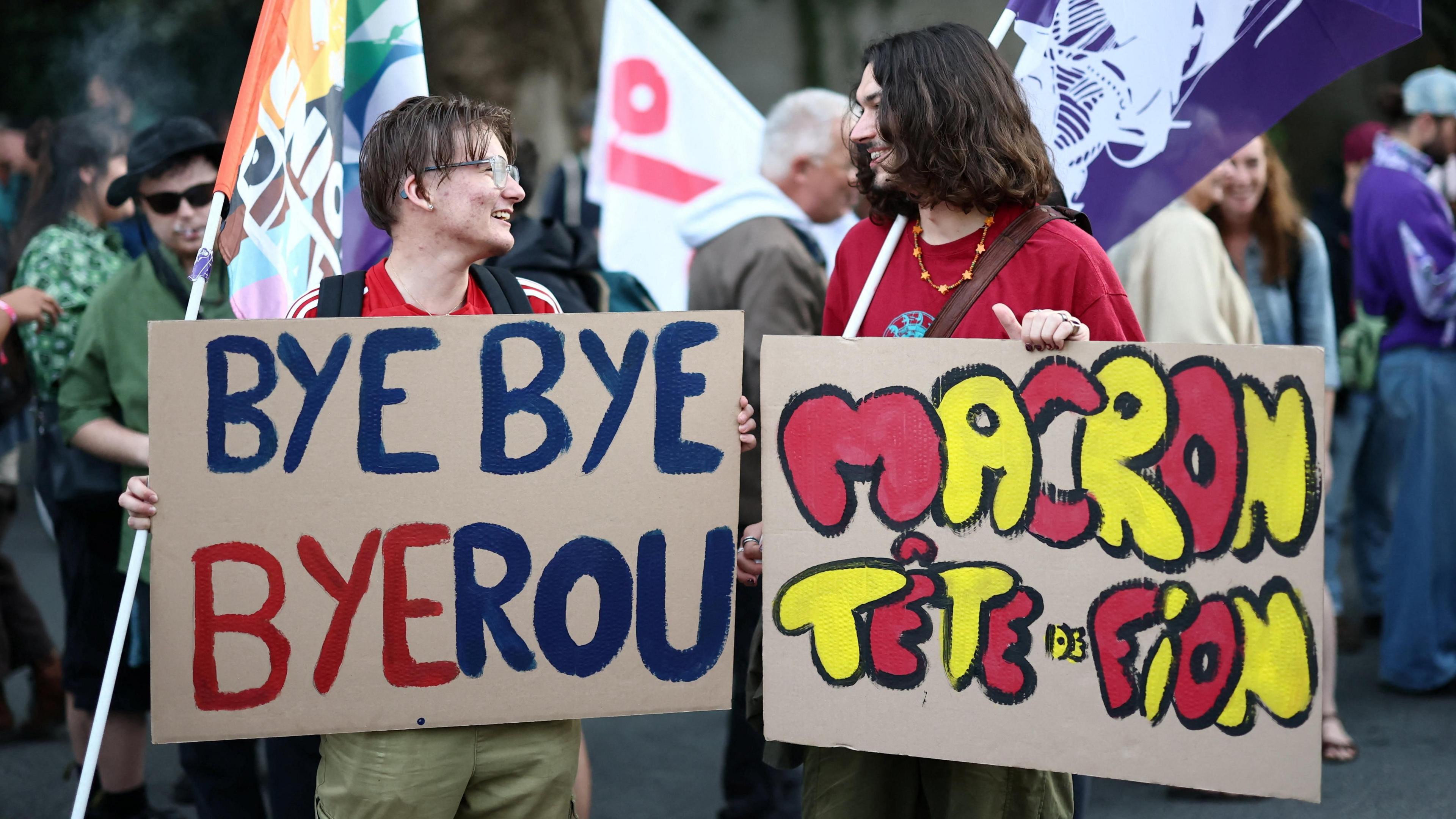 Two people look at eachother holding colourful signs that say "Bye bye Byerou" and "Macron Tete de Fion".