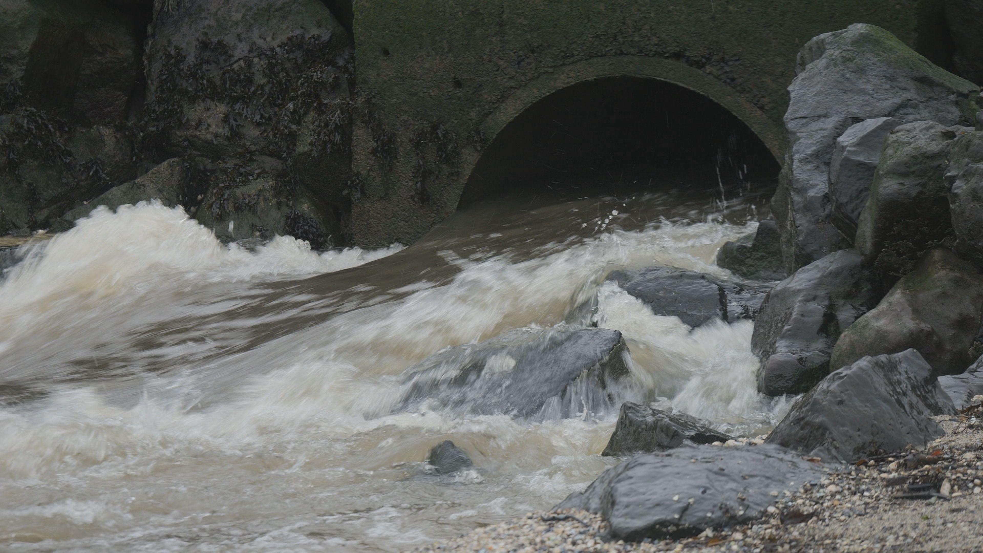 A large circular storm drain, approximately three feet in diamater, has lots of brown water gushing out of it onto a beach. There are lots of smaller rocks built up to the sides of the storm overflow.