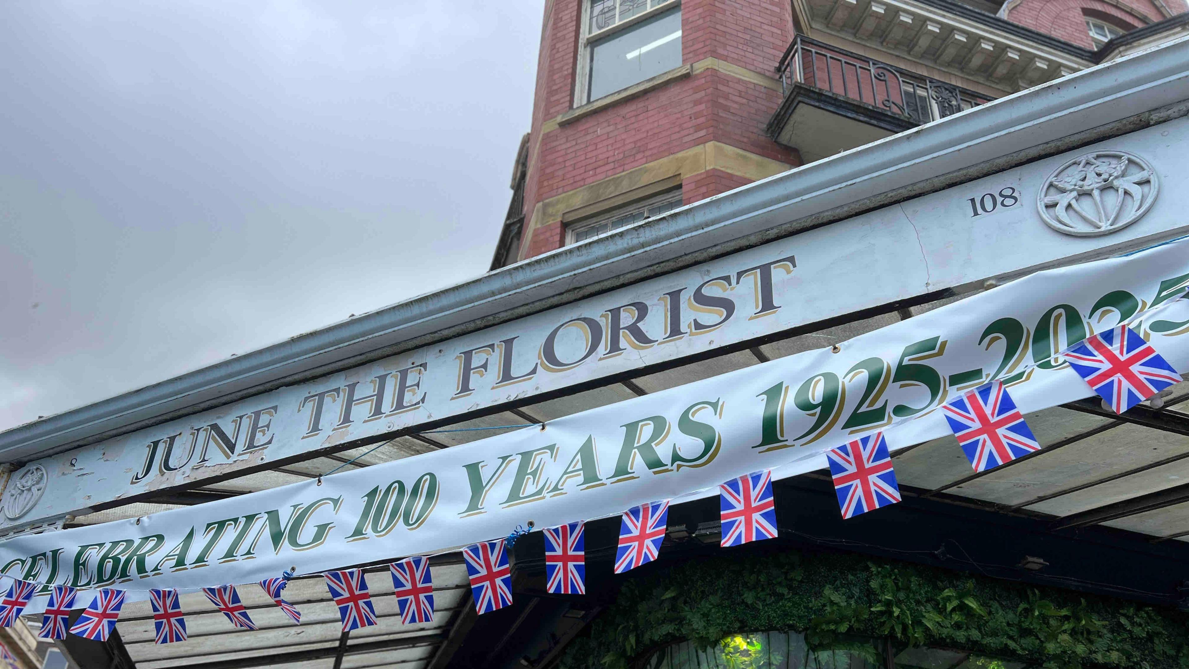 A close up shot of the June the Florist shop sign, a banner reads "celebrating 100 years 1025-2025". There is Union Jack bunting below the banner.