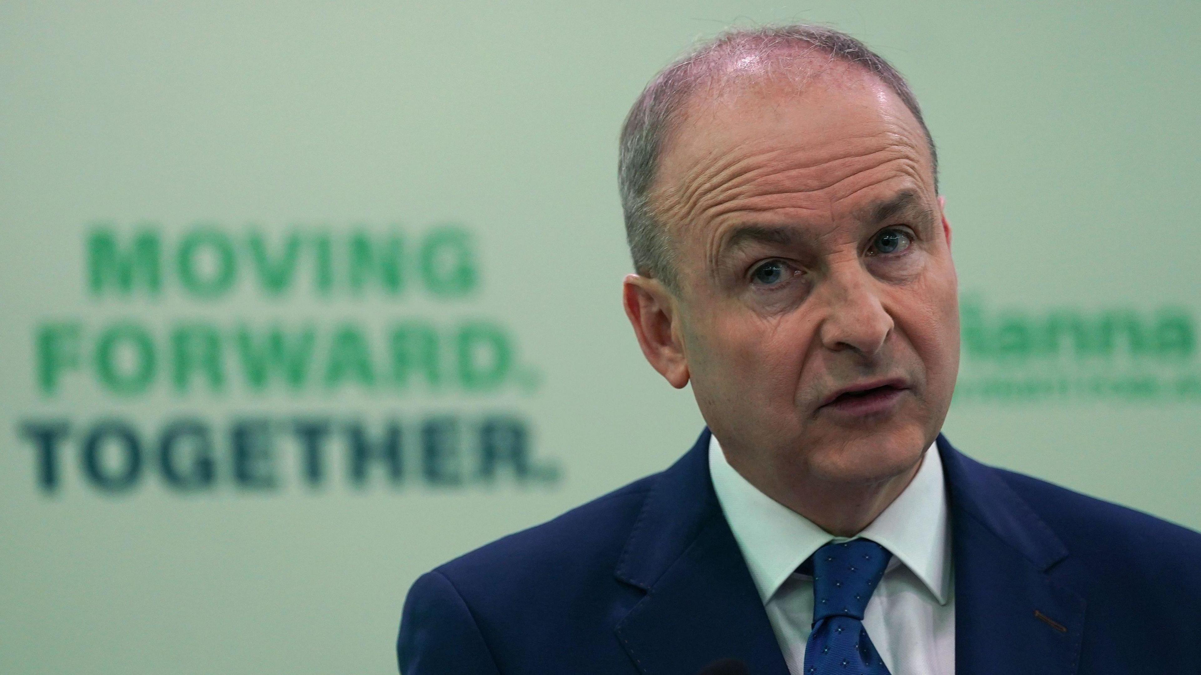 A man in a navy blue suit, he is stood in front of a sign which says 'moving forward, together'. 