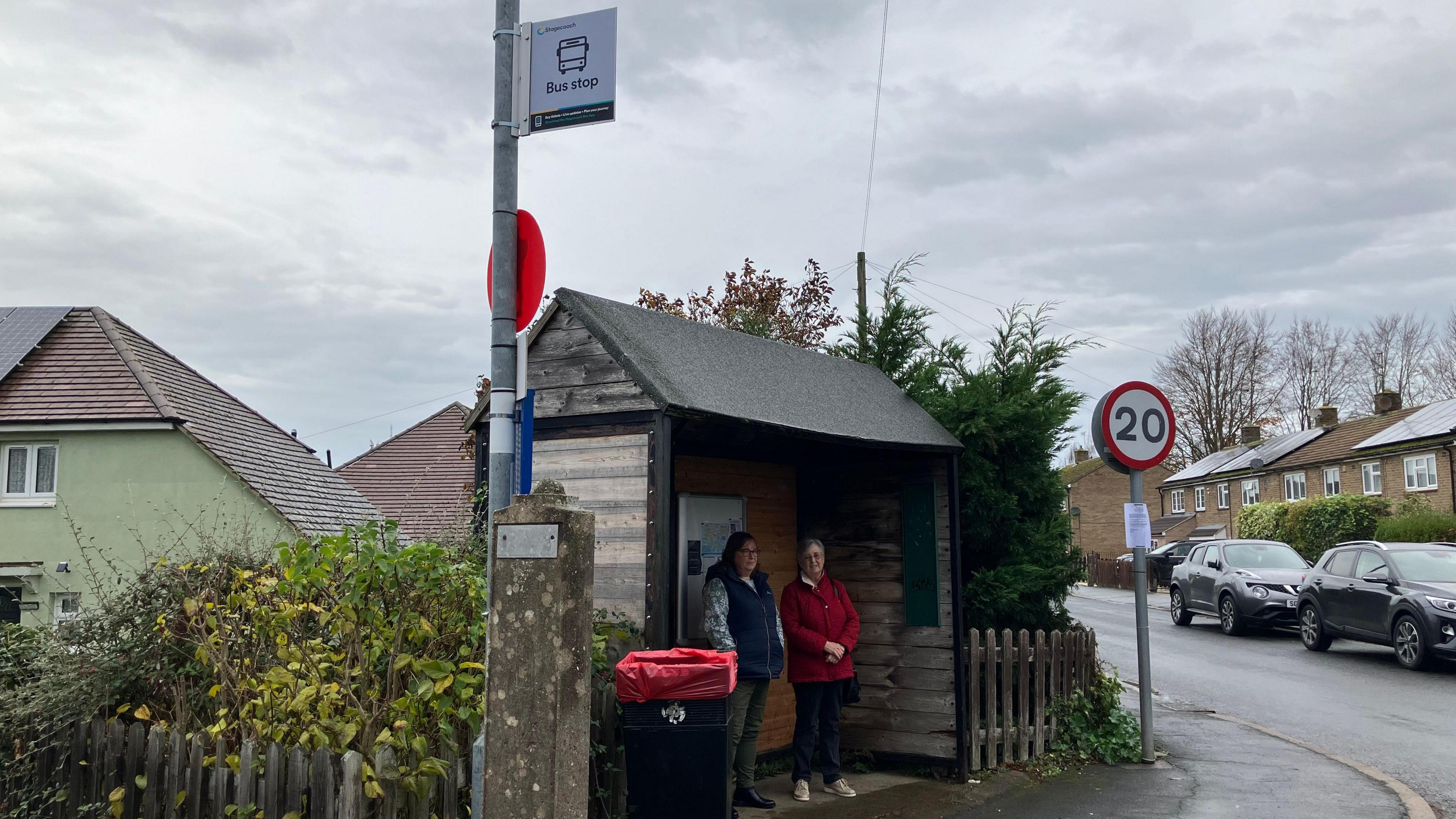 A wooden bus shelter with two women standing inside it. It is a cloudy day and it has been raining. There are houses to the right and left of the frame.