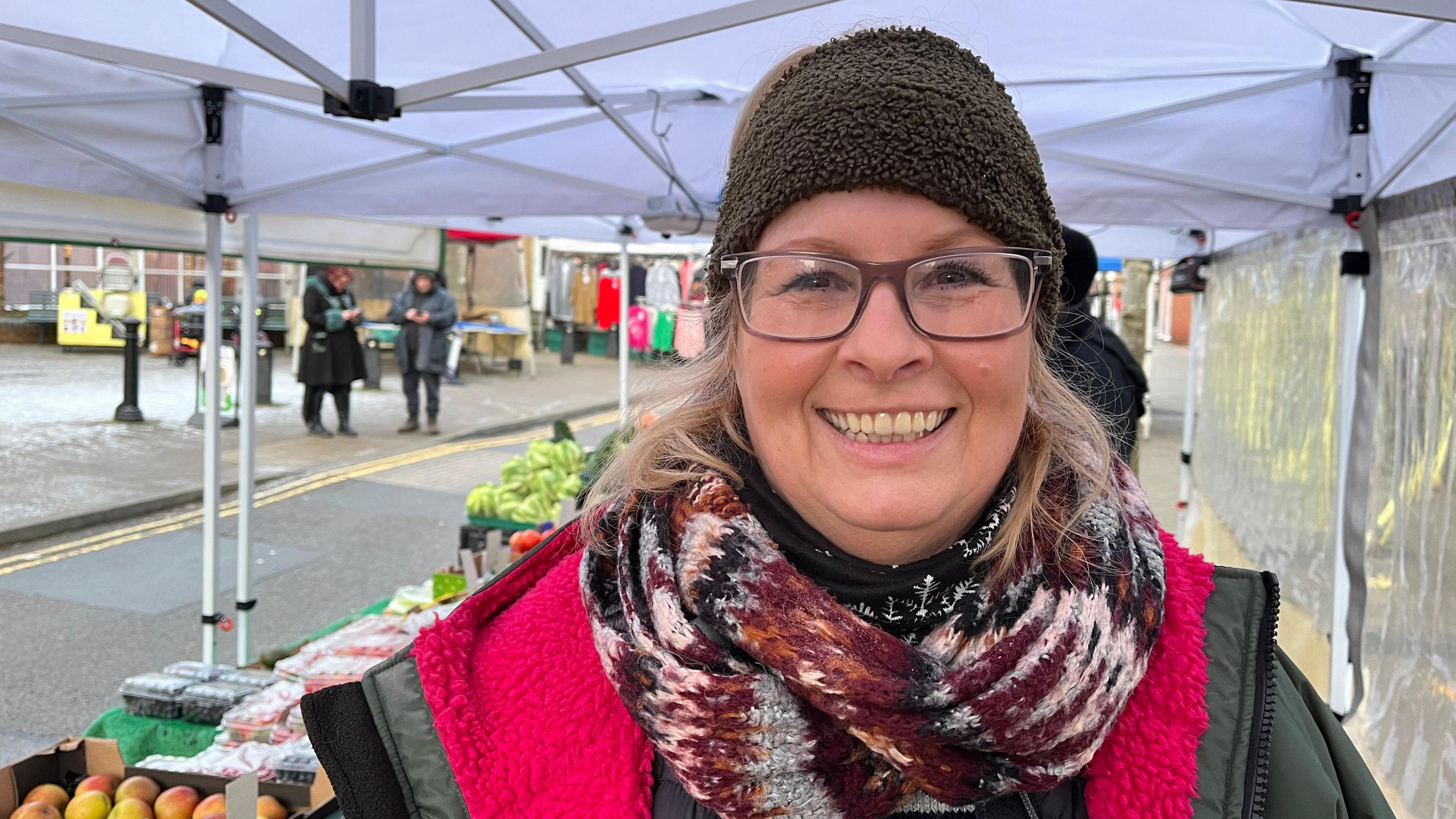 Jan looking into the camera and smiling, she's wearing a winter coat, hat and scarf. Behind her you can see fruits and vegetables laid out on her stall, with a white gazebo covering it. 