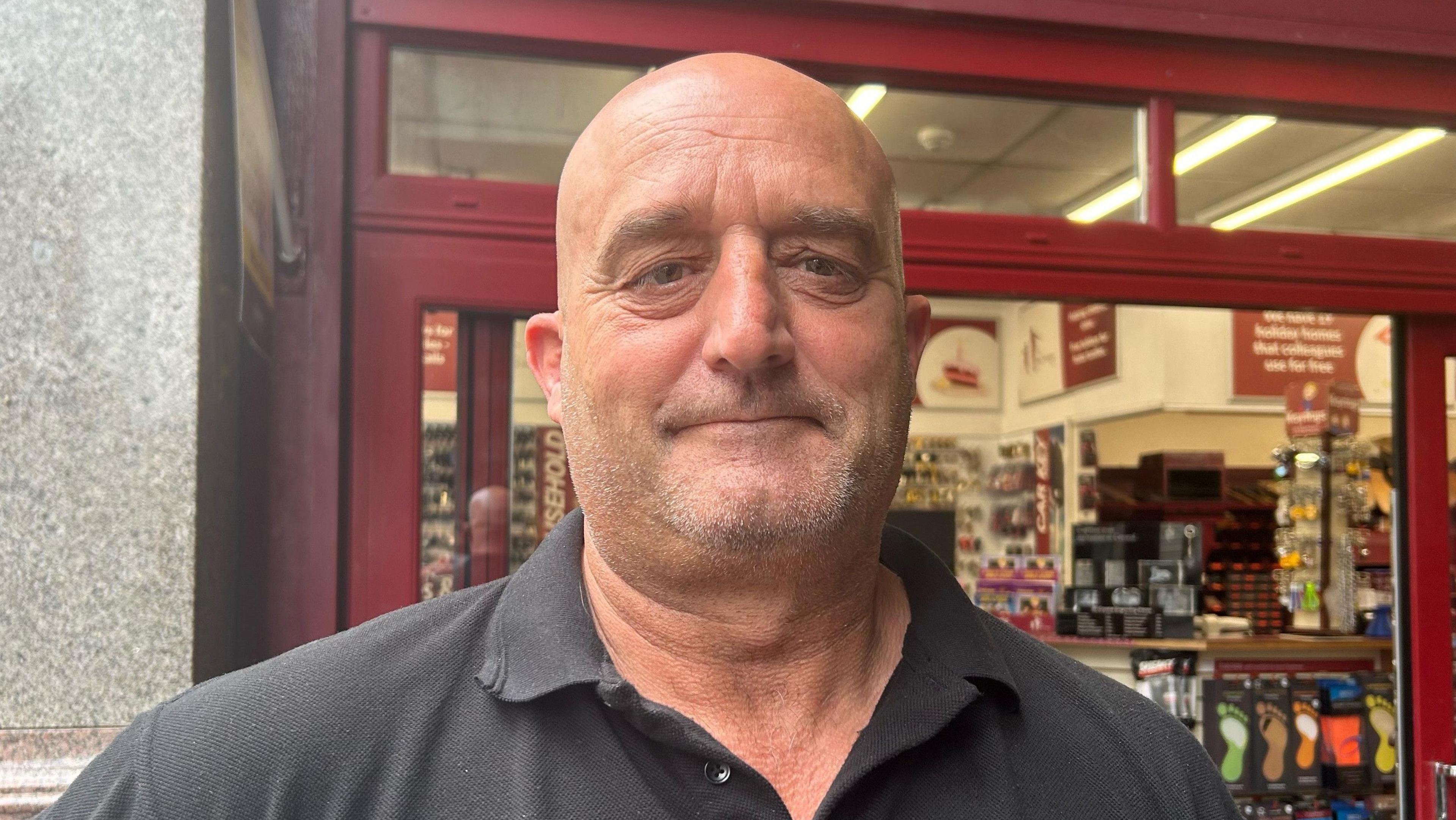A man with light stubble is looking at the camera. He is wearing a black polo shirt and is standing outside a shop.