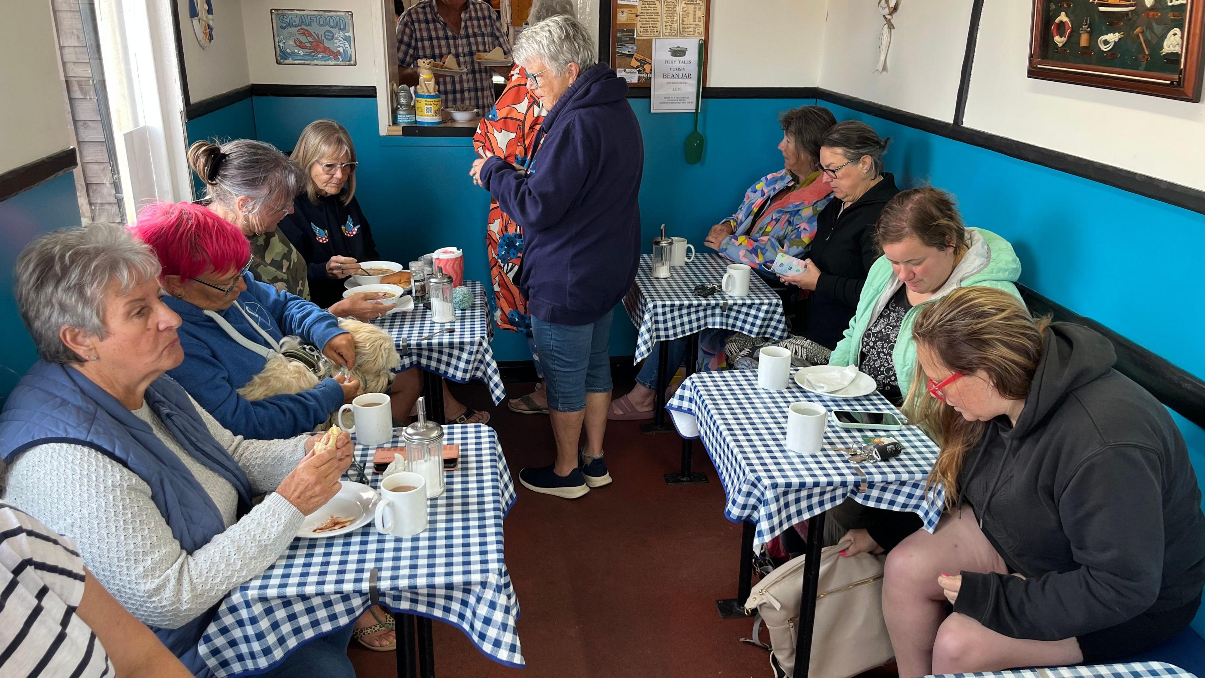 A group of women sitting at a number of tables having a breakfast of bean jar, bacon sandwiches and baps. There is a dog on one of the women's laps.
