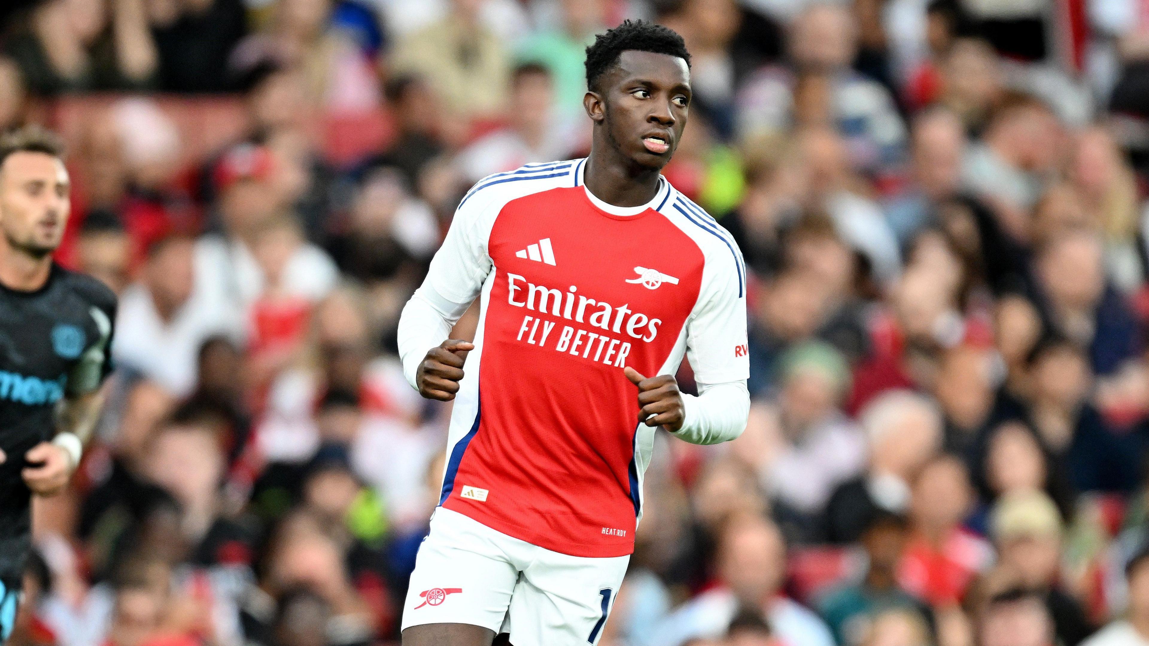 Eddie Nketiah, wearing Arsenal's home red kit with white sleeves, has his arms partially out with his fists closed as he appears to jog. There are fans in the stand who are in the background but are blurred.