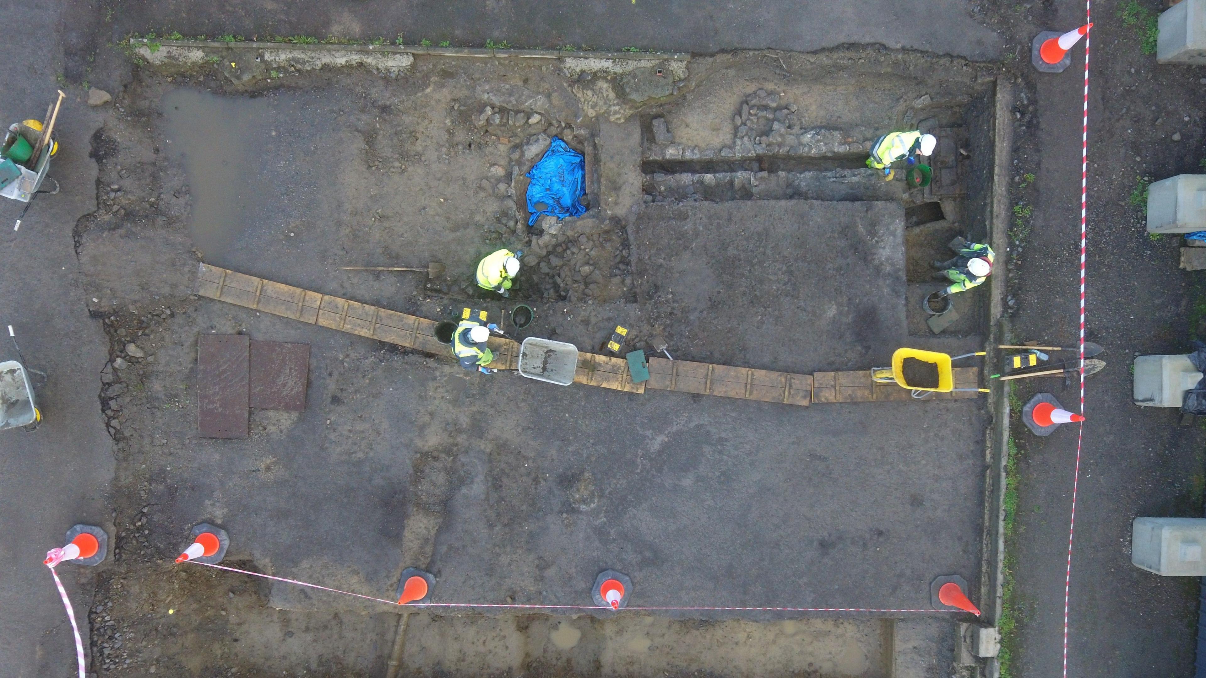 Image from above of excavators working at a site. They are wearing hard hats and high vis jackets. There is a cordon around the site formed by traffic cones and red and white striped tape.