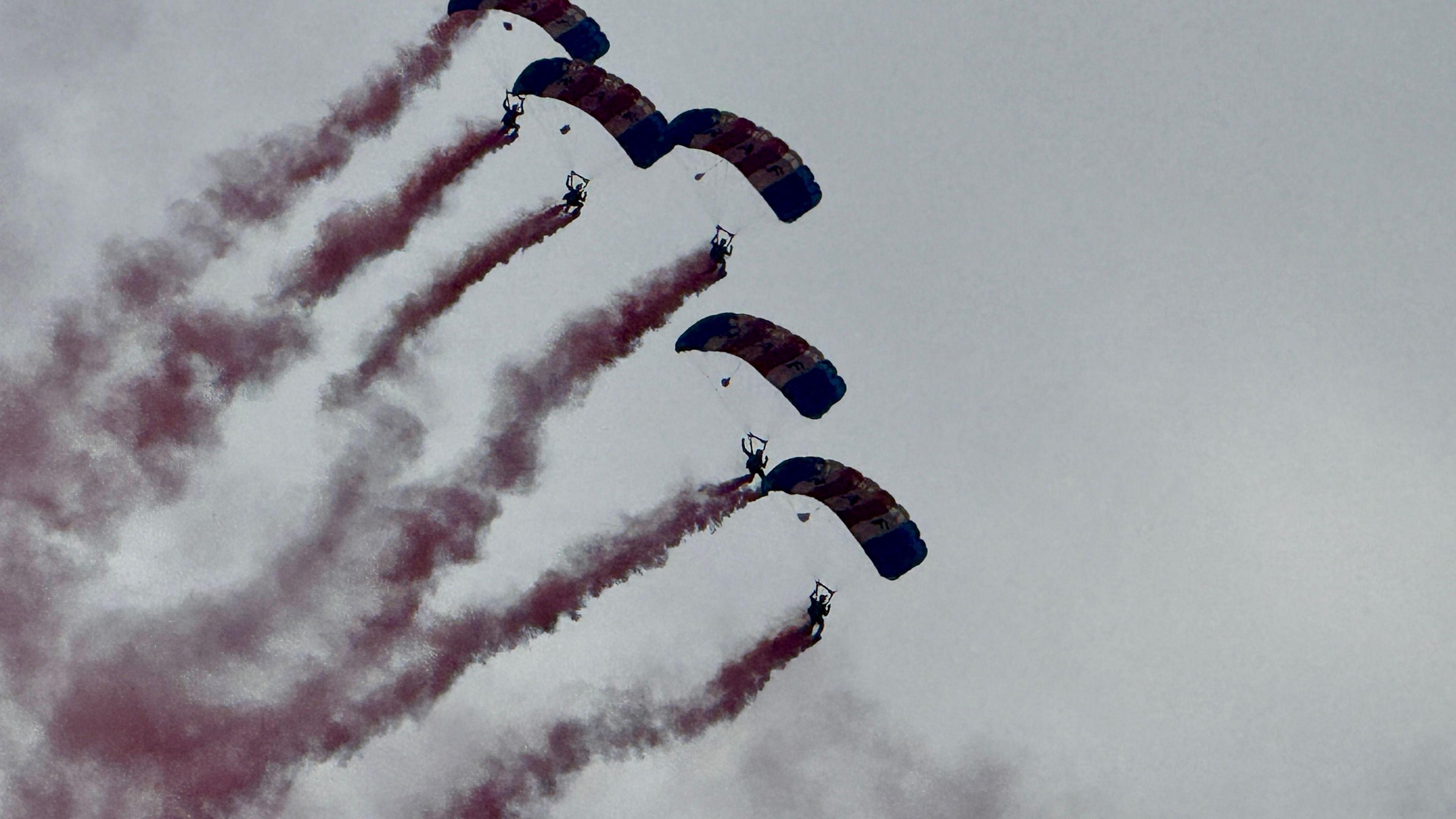 Five people with parachutes deployed falling from a grey sky as red-coloured smoke trails behind each of them.
