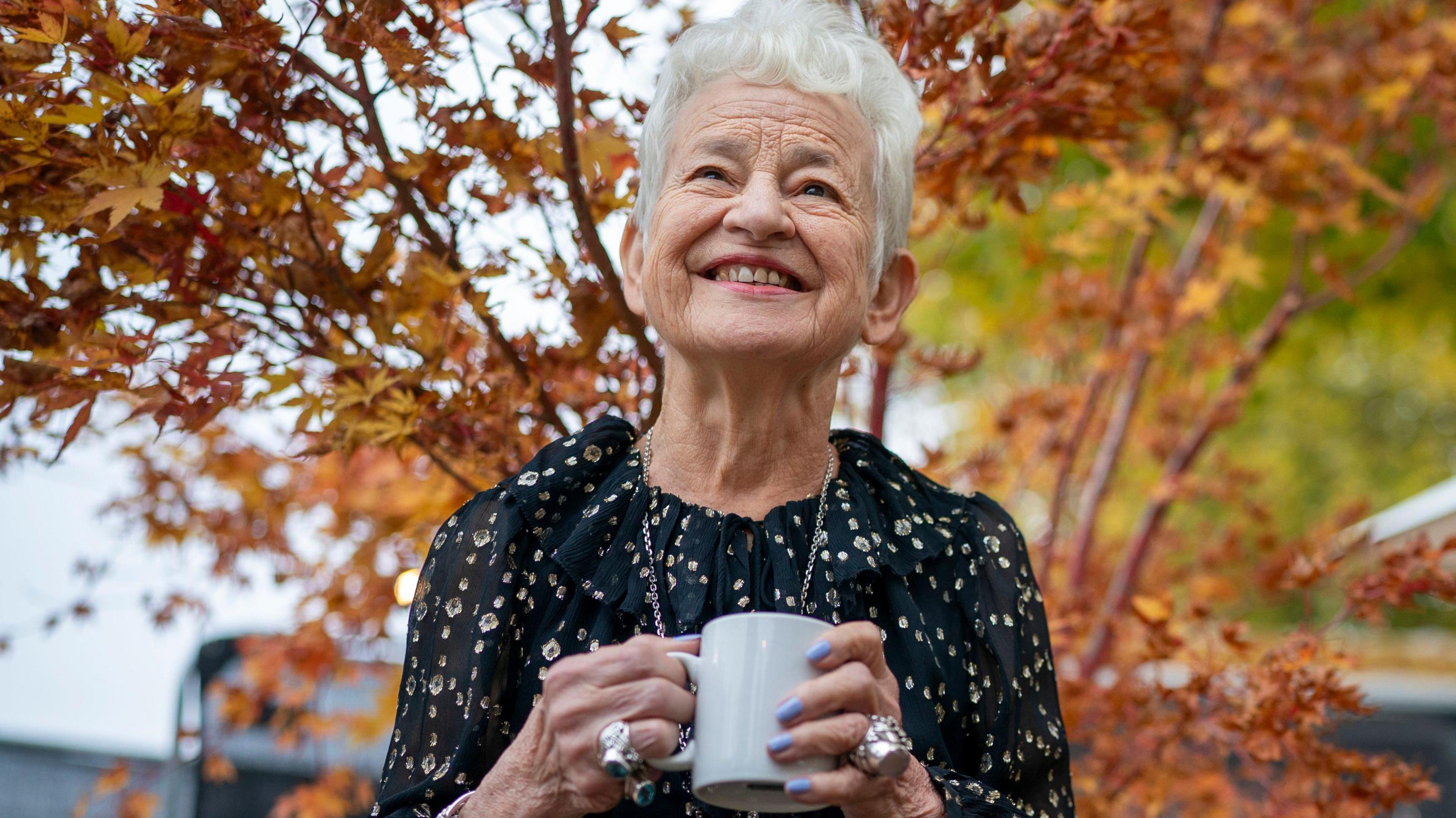 Jacqueline Wilson, the children's author, smiles as she stands in front of a browning tree, clutching a mug. She has short white hair, a black top with a ruffled neck and flower detailing, lots of chunky silver rings, and pastel blue painted nails.