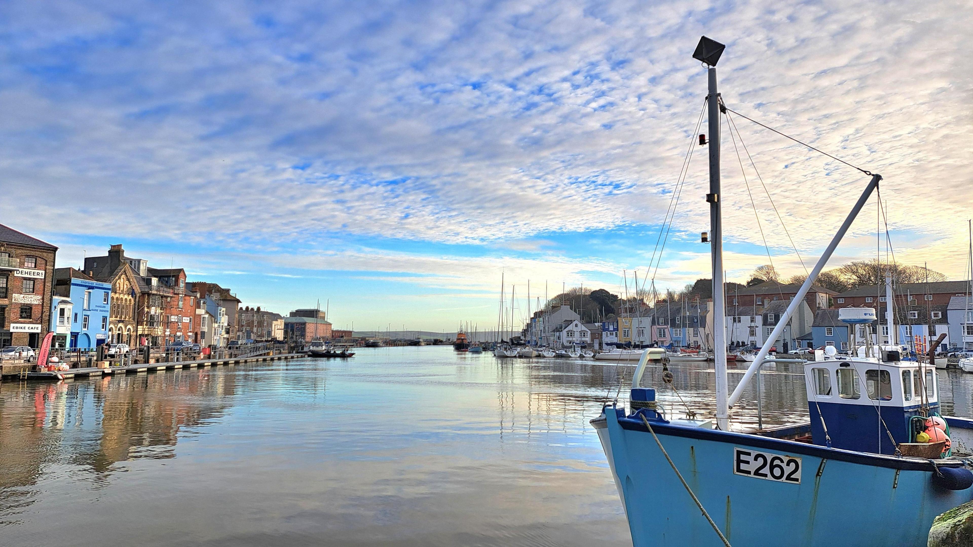 Weymouth Harbour under a clear blue sky on a sunny day. There is a blue fishing boat in the foreground. The harbour is lined by buildings and the sky is reflected in the water.