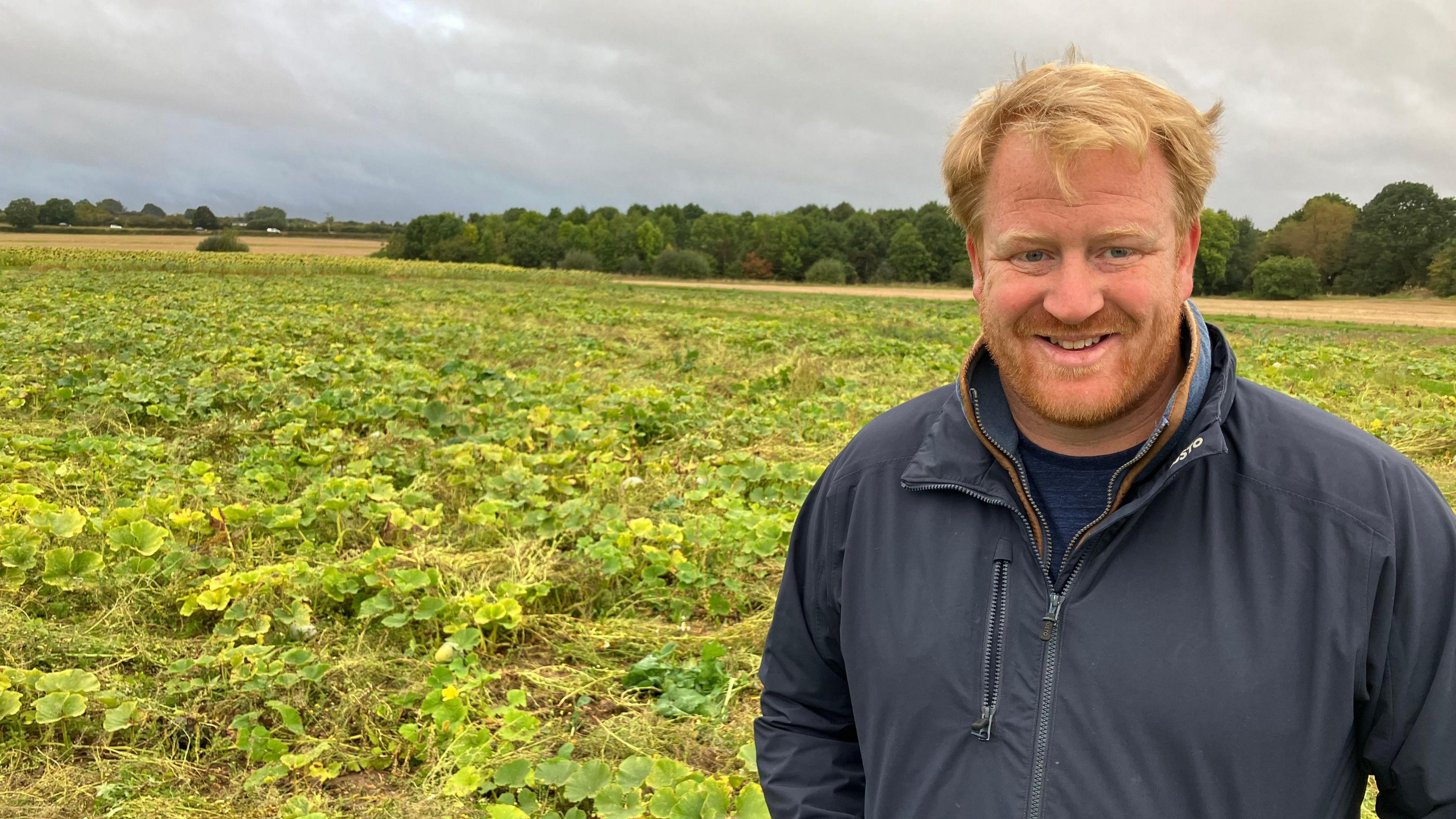 A man in a navy jacket with blonde hair stands in a large squash field.