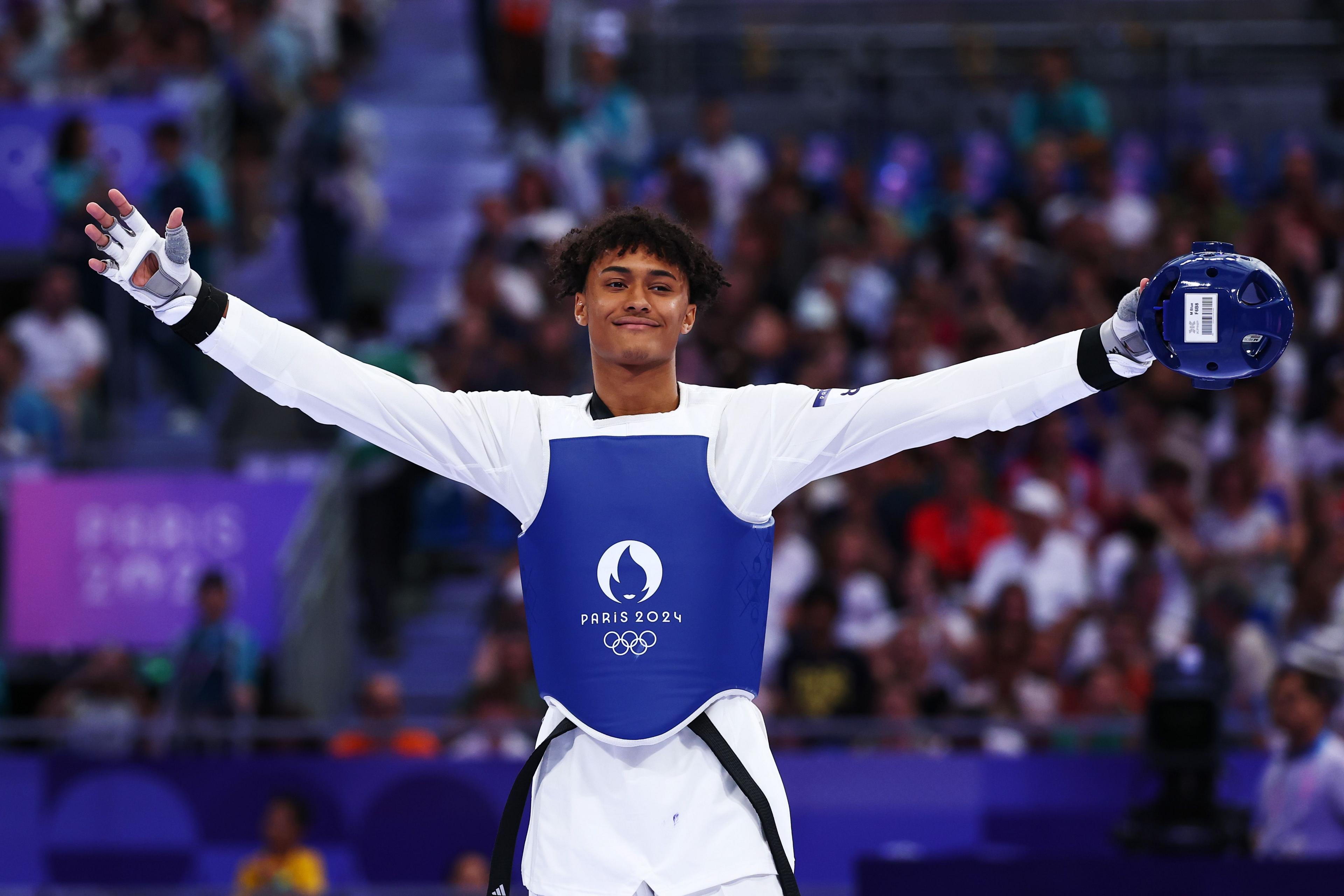 Caden Cunningham stands with his arms stretched wide, a blue helmet in one hand. He is smiling and wearing all white with a blue vest with the Paris 2024 logo on. The crowd behind him is blurred.