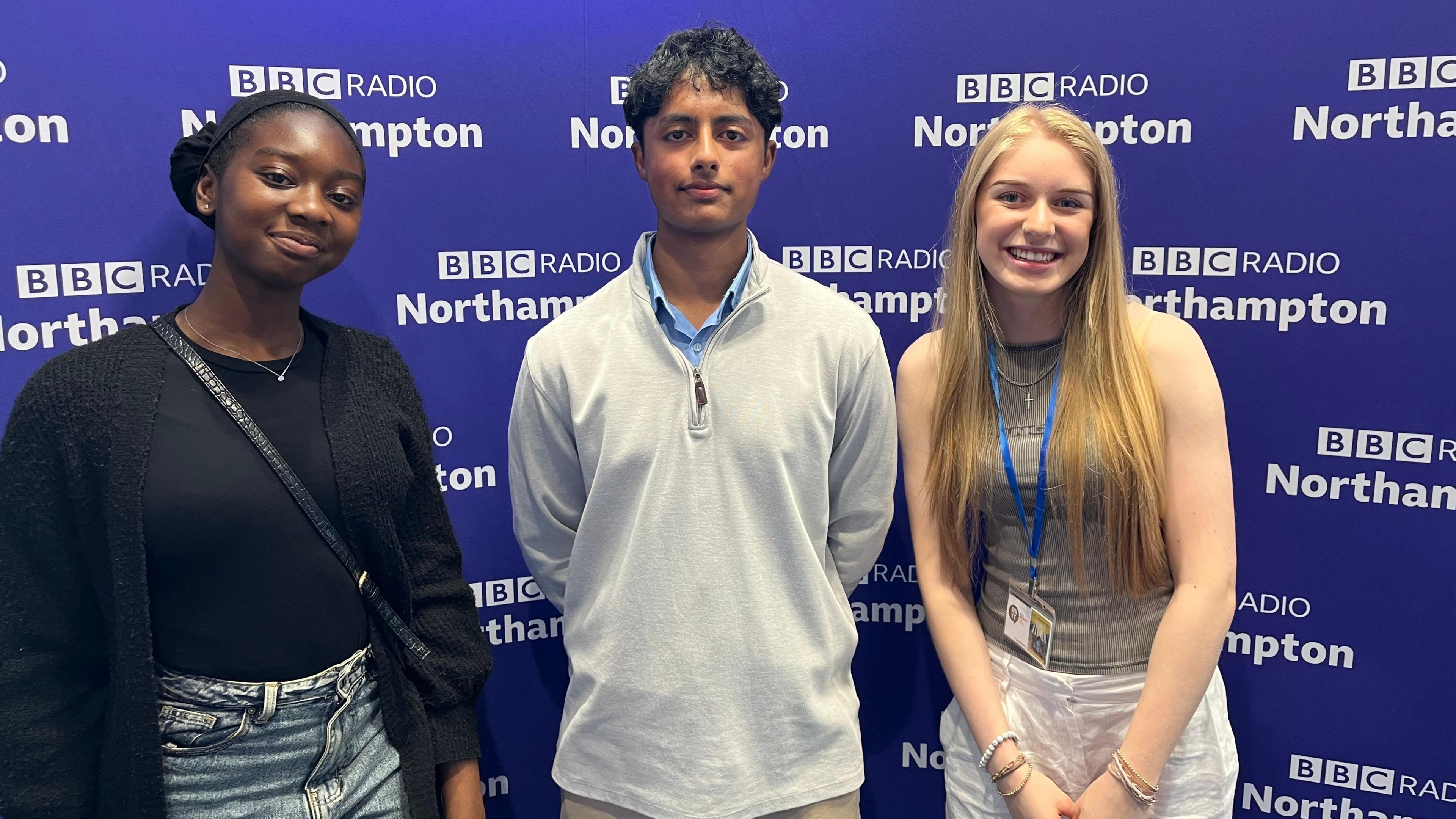 Two teenage girls and a teenage boy stand in front of a purple backdrop 