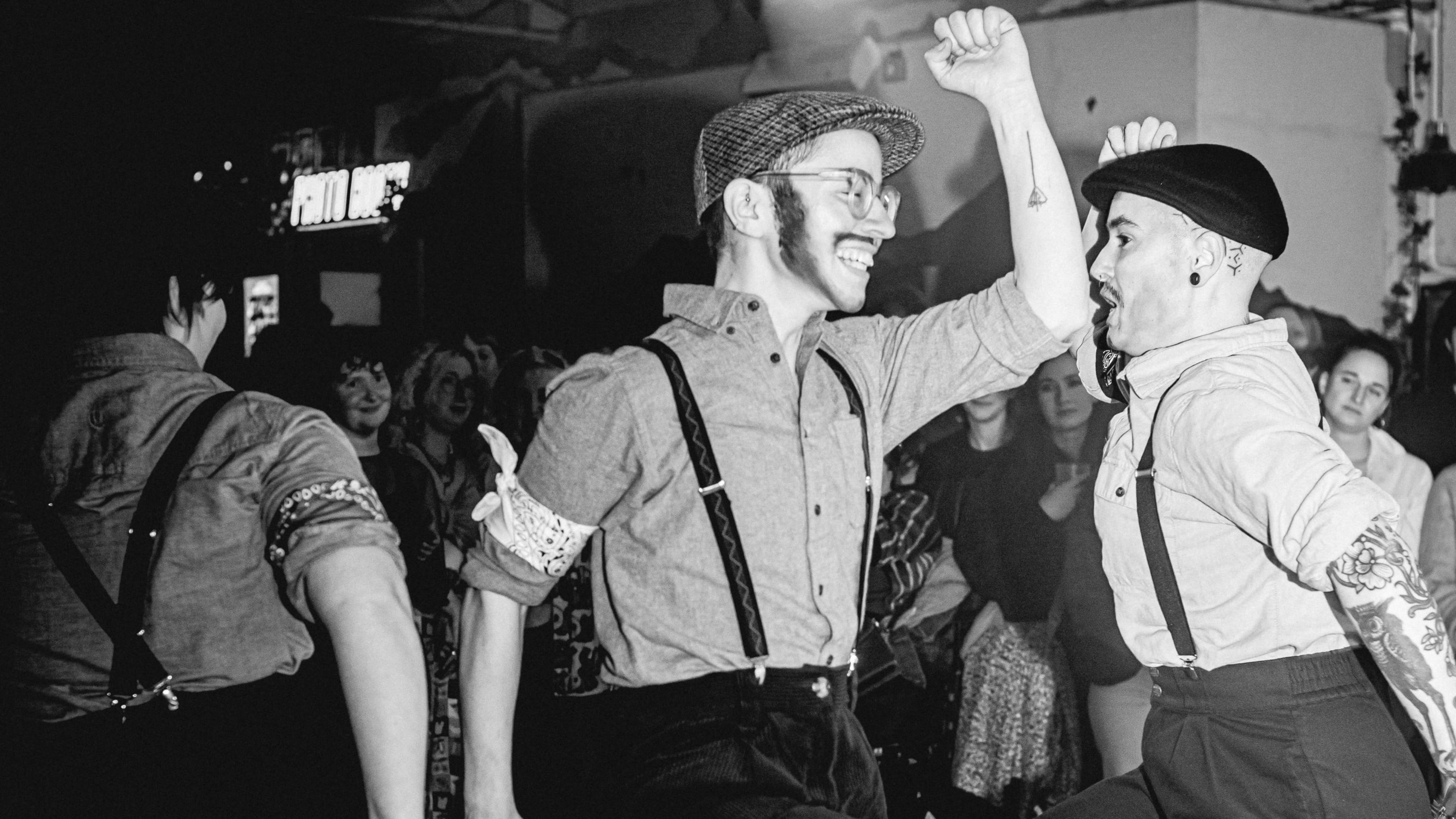 Black and white photo showing members of Molly No-mates dancing inside at a venue wearing old-fashioned caps, trousers and braces.